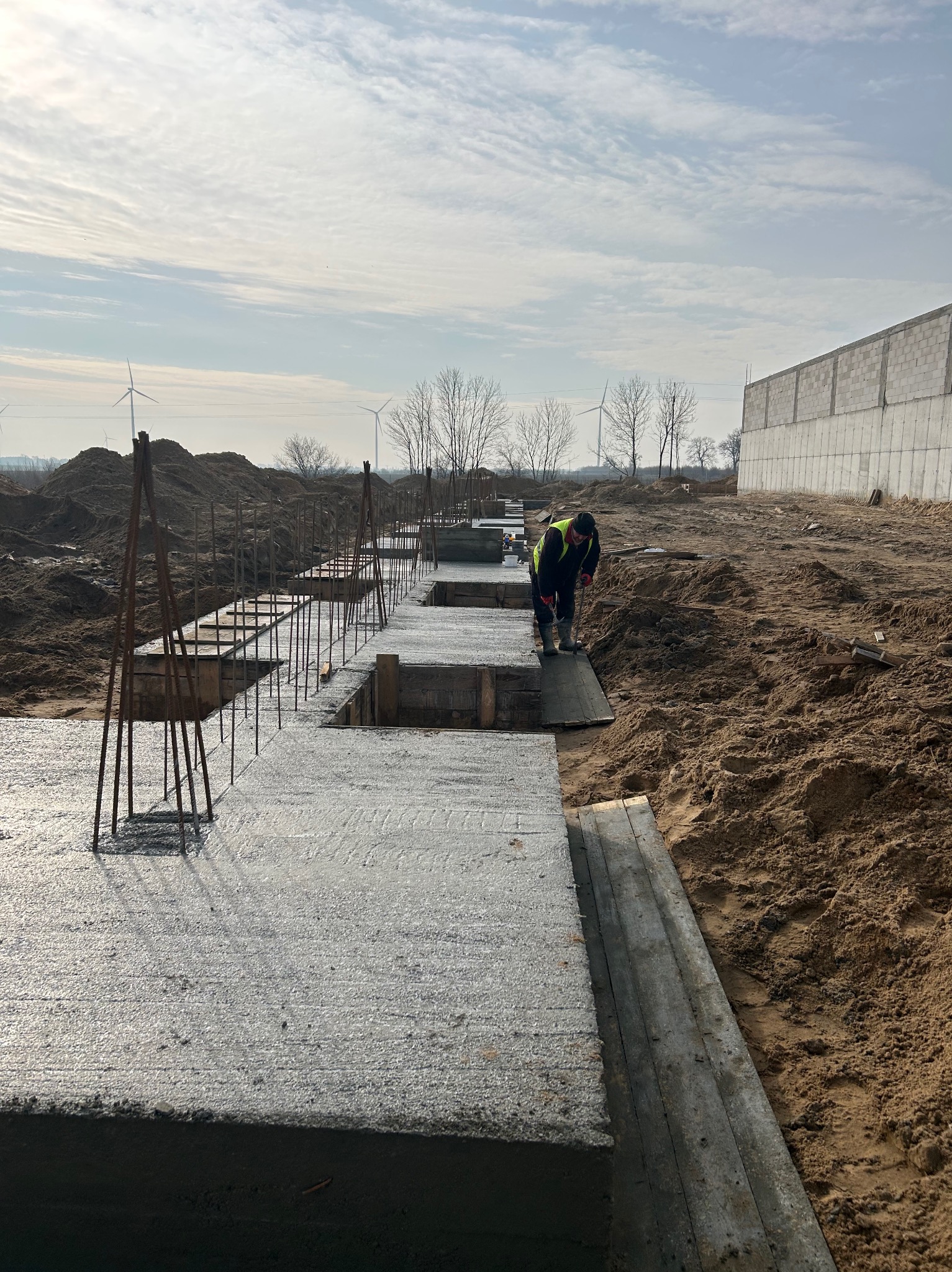 Zalane fundamenty pod budowę. Widoczne szalunki, wystające pręty zbrojeniowe, pracownik w odblaskowej kamizelce. W tle widać turbiny wiatrowe i budynek w trakcie budowy.