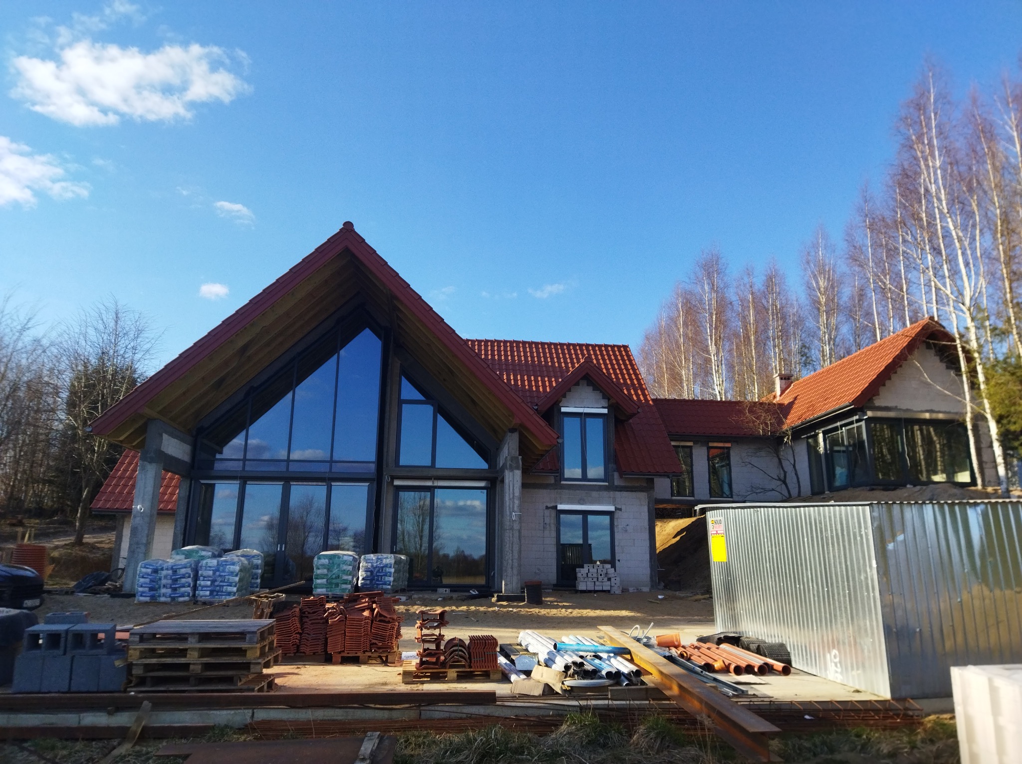 Budowa domu w stanie surowym otwartym z widocznymi materiałami budowlanymi, czerwona dachówka, duże okna z refleksami nieba. W tle drzewa i błękitne niebo.