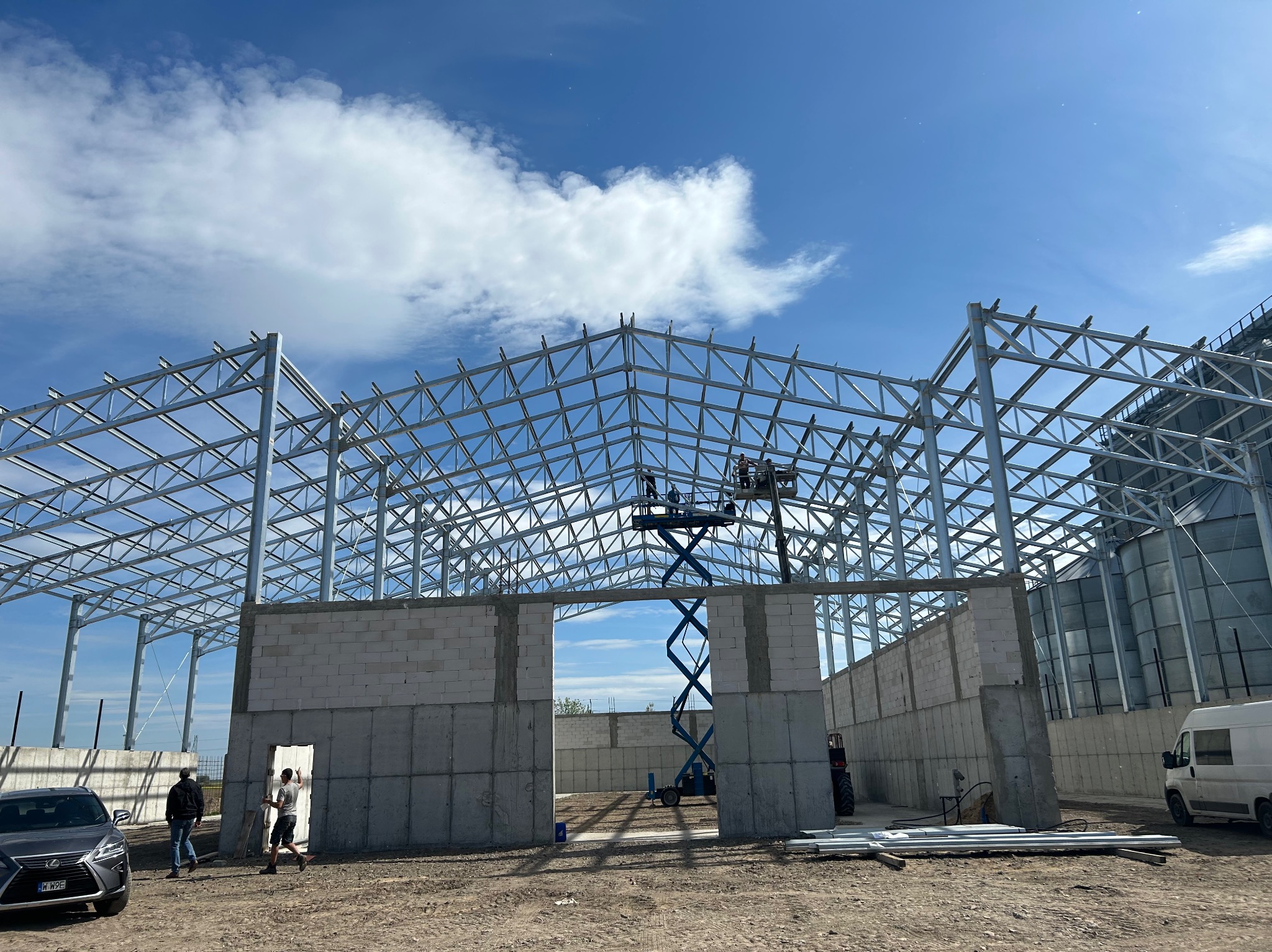 Stalowa konstrukcja budynku w trakcie budowy z widocznymi fragmentami ścian z betonu komórkowego, podnośnikiem koszowym i pracownikami na zewnątrz.