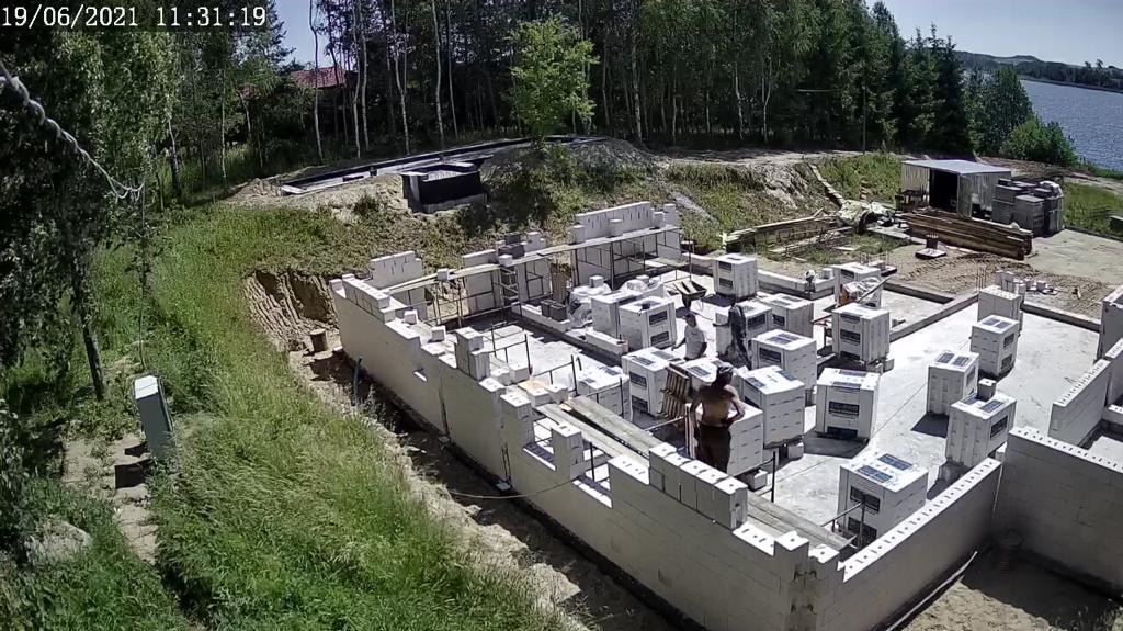Budowa fundamentów domu z bloczków betonowych na działce z widokiem na jezioro, w tle las. Widoczni pracownicy budowlani w trakcie pracy.