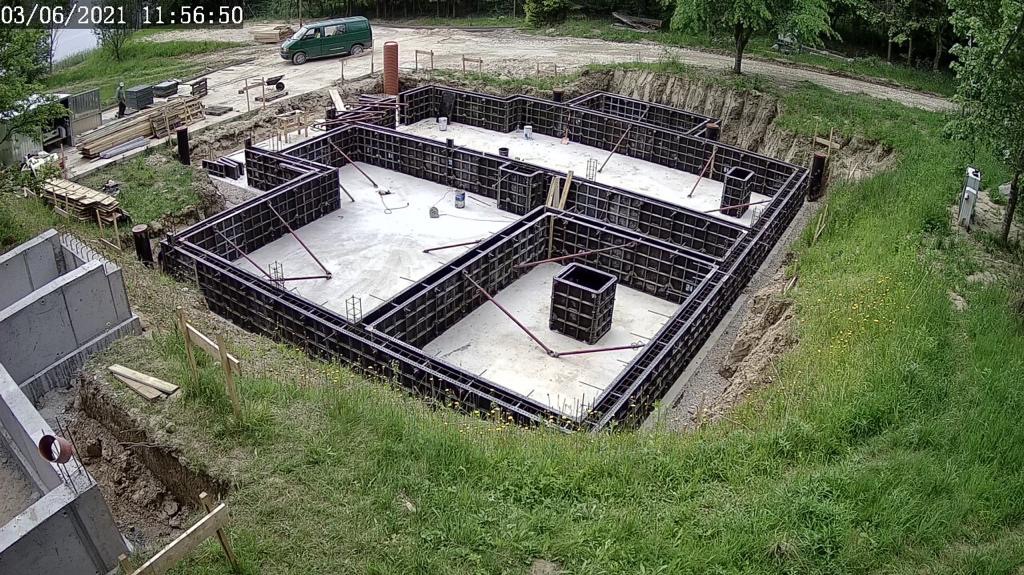 Fundamenty domu w trakcie budowy, z widocznym zbrojeniem i szalunkiem. Ujęcie z góry, w tle zielony krajobraz i częściowo widoczny samochód dostawczy.