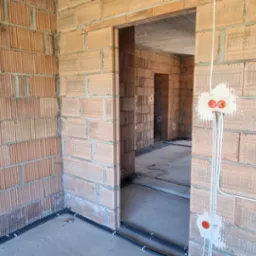 Surowy stan budynku z cegły z widoczną instalacją elektryczną: kable w peszlach ułożone przy podłodze i puszki elektryczne zamontowane w ścianie.