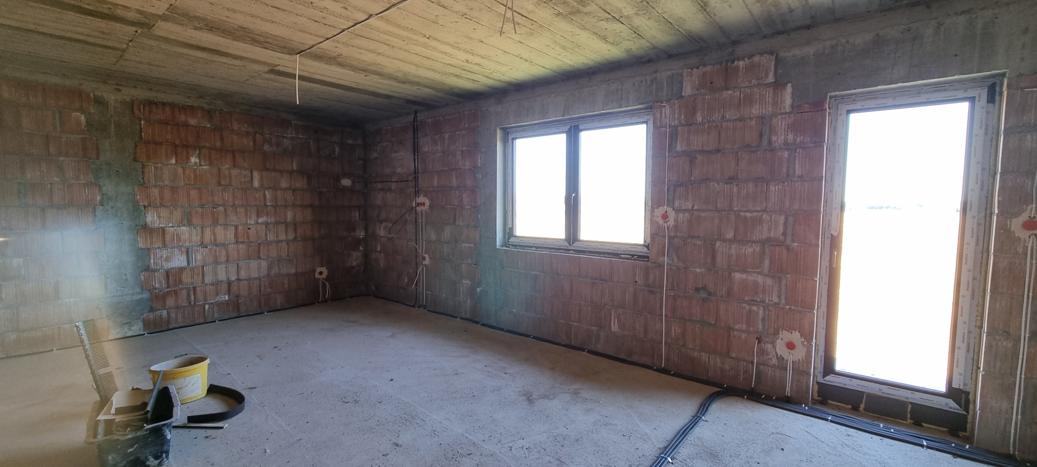 Surowy stan wnętrza z instalacją elektryczną: widoczne przewody, puszki, ceglane ściany, okno i drzwi balkonowe. Na podłodze leżą narzędzia i wiadro.