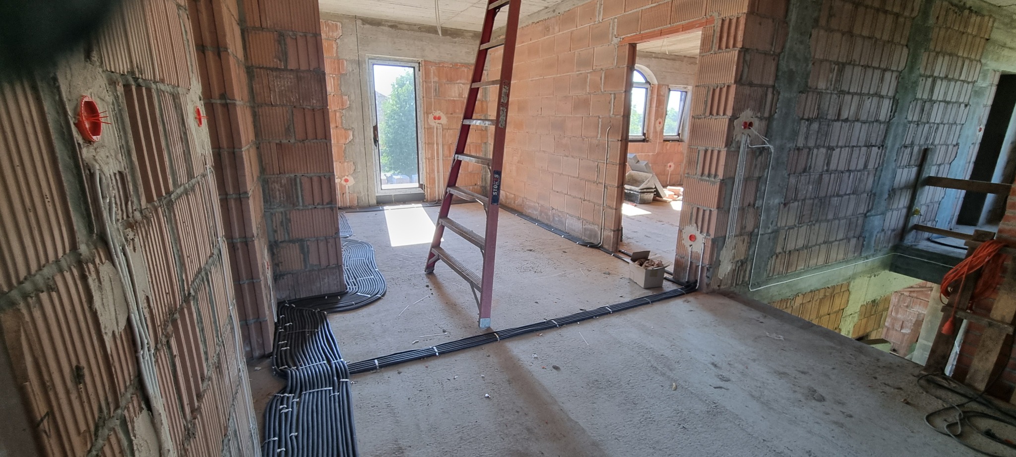 Instalacja elektryczna w trakcie budowy: kable ułożone na podłodze, puszki elektryczne w ścianach z cegły. Widoczna drabina i surowy stan wykończenia wnętrza.