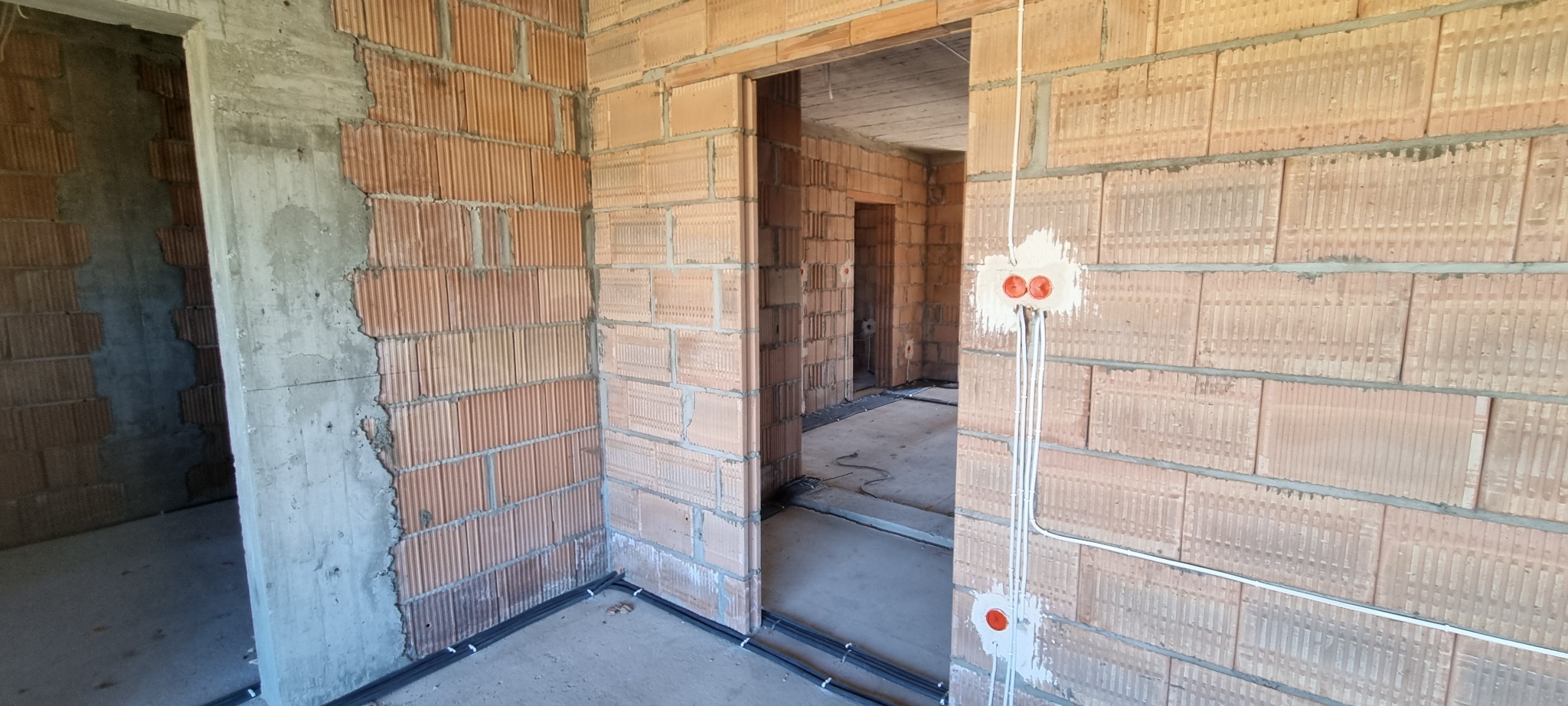 Surowy stan budynku z cegły z widoczną instalacją elektryczną: kable w peszlach ułożone przy podłodze i puszki elektryczne zamontowane w ścianie.