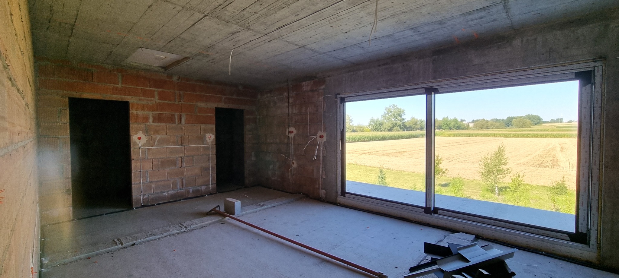 Surowy stan wnętrza z widocznymi instalacjami elektrycznymi w ścianach z cegły, betonowym stropem i dużym oknem z widokiem na pola. Wnętrze w trakcie prac wykończeniowych.