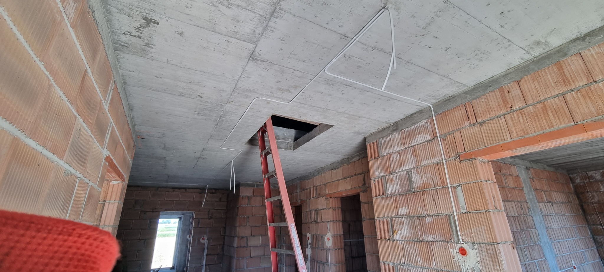 Surowy strop z instalacją elektryczną w rurkach, czerwona drabina w otworze. Ściany z czerwonej cegły dziurawki. W tle widok na zewnątrz przez okno.