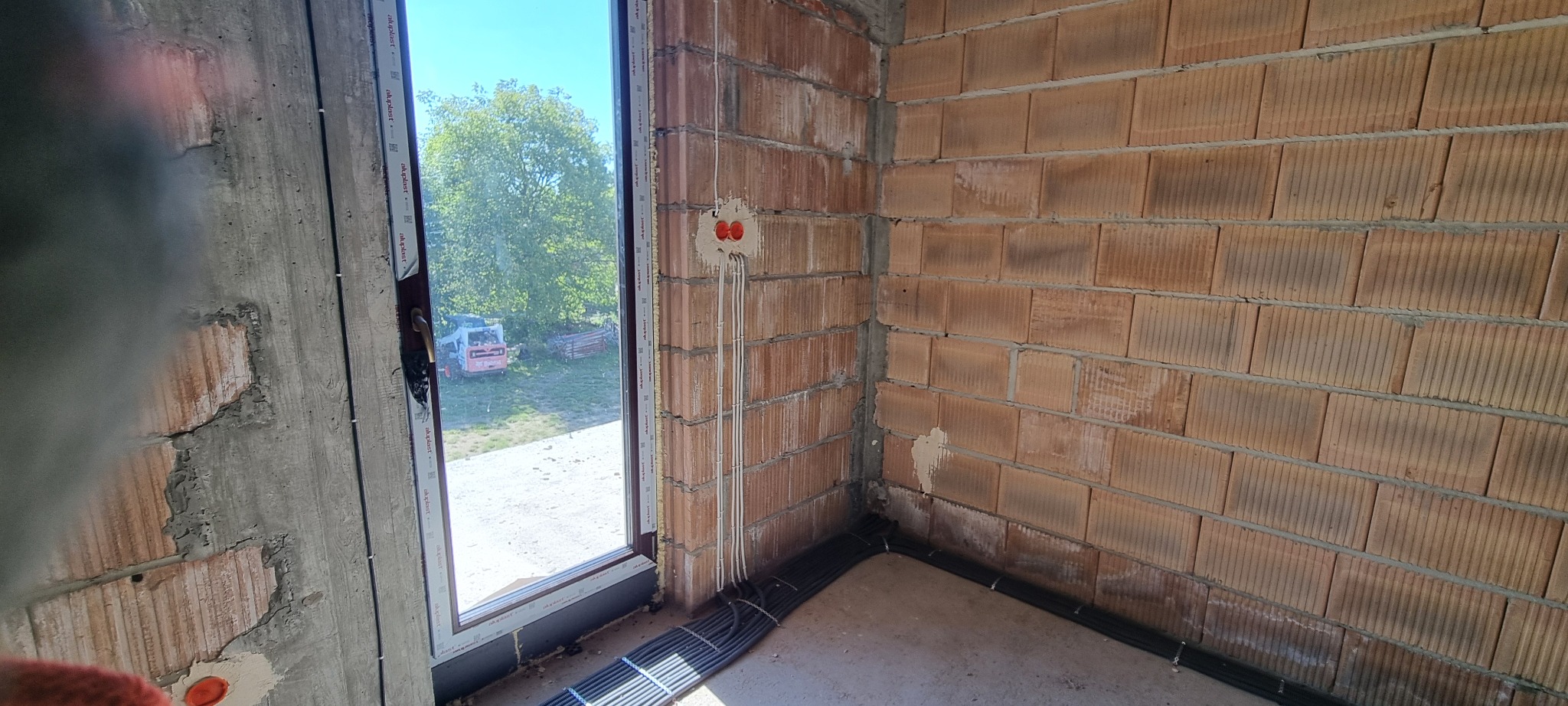 Instalacja elektryczna w surowym budynku z czerwonej cegły w Charbinowicach. Widoczne kable i puszki elektryczne przy oknie. Prace elektryczne w trakcie realizacji.