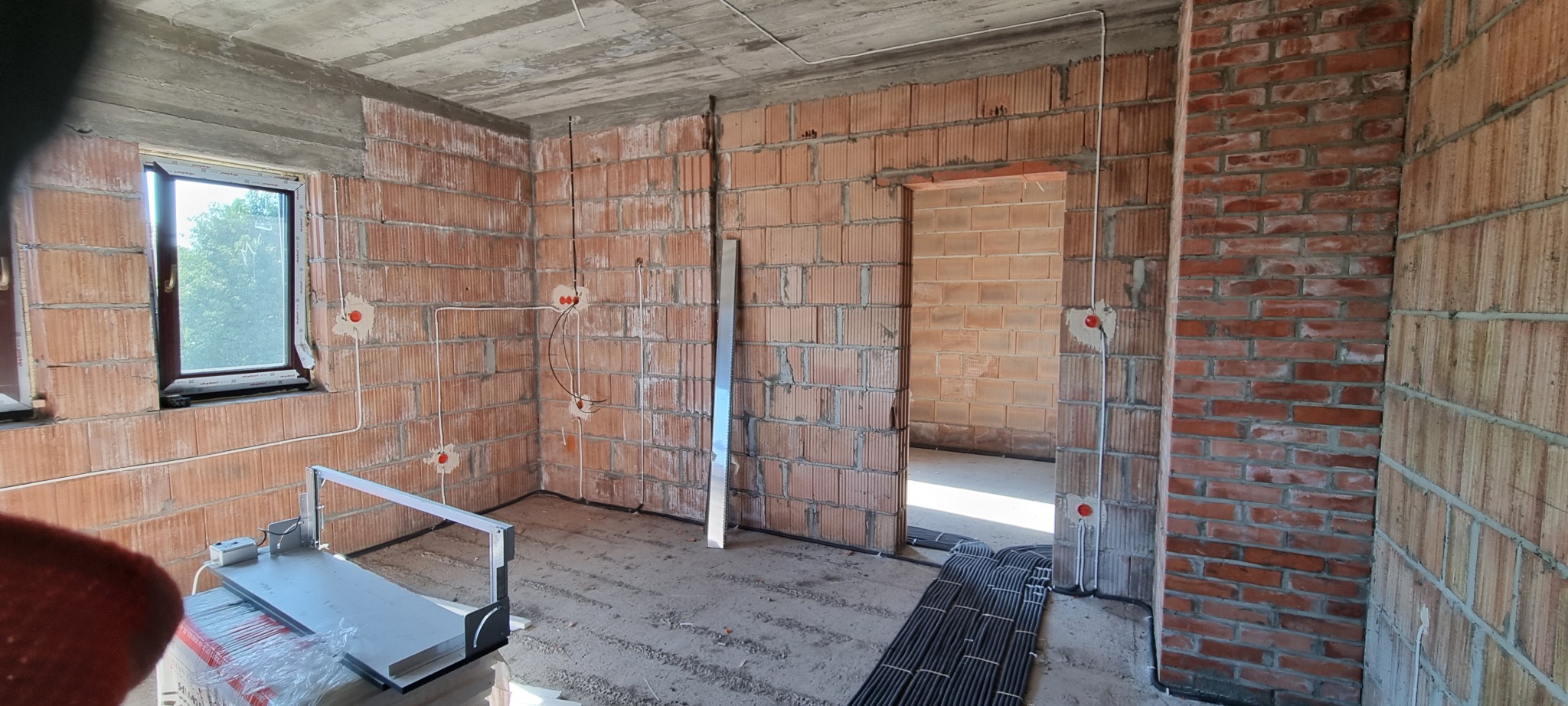 Wnętrze w trakcie prac: ściany z cegły, okno, instalacja elektryczna w peszlach. Widoczne kable na podłodze i puszki elektryczne w ścianach. Surowy stan wykończenia.