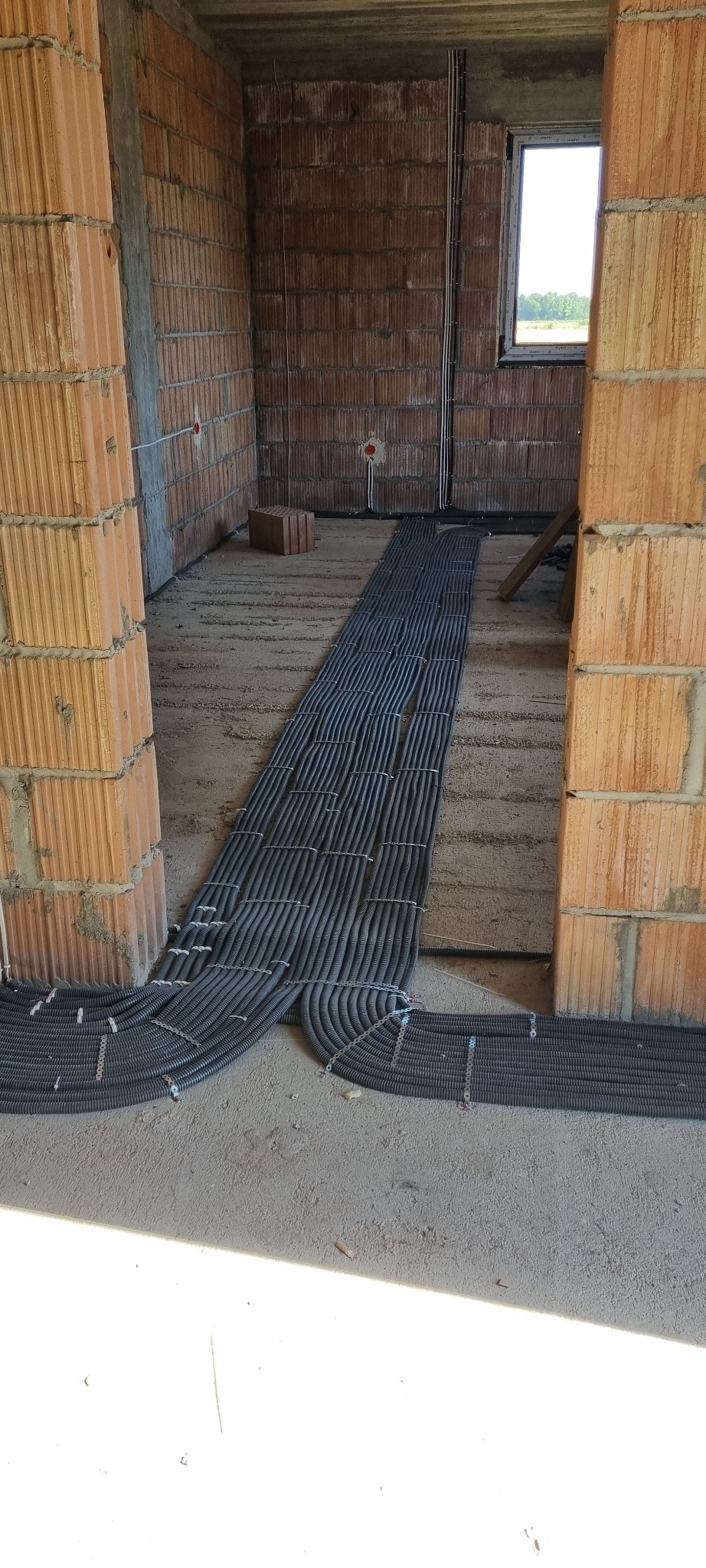 Wnętrze w trakcie budowy z widocznymi, ułożonymi w rzędach, czarnymi rurami instalacji elektrycznej na podłodze, przymocowanymi metalowymi uchwytami do podłoża.