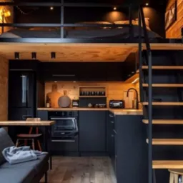 Małe, ale funkcjonalne wnętrze z antresolą sypialnianą, czarną zabudową kuchenną na tle drewnianej ściany, drewnianymi schodami i szarą sofą, oświetlone punktowymi lampami.
