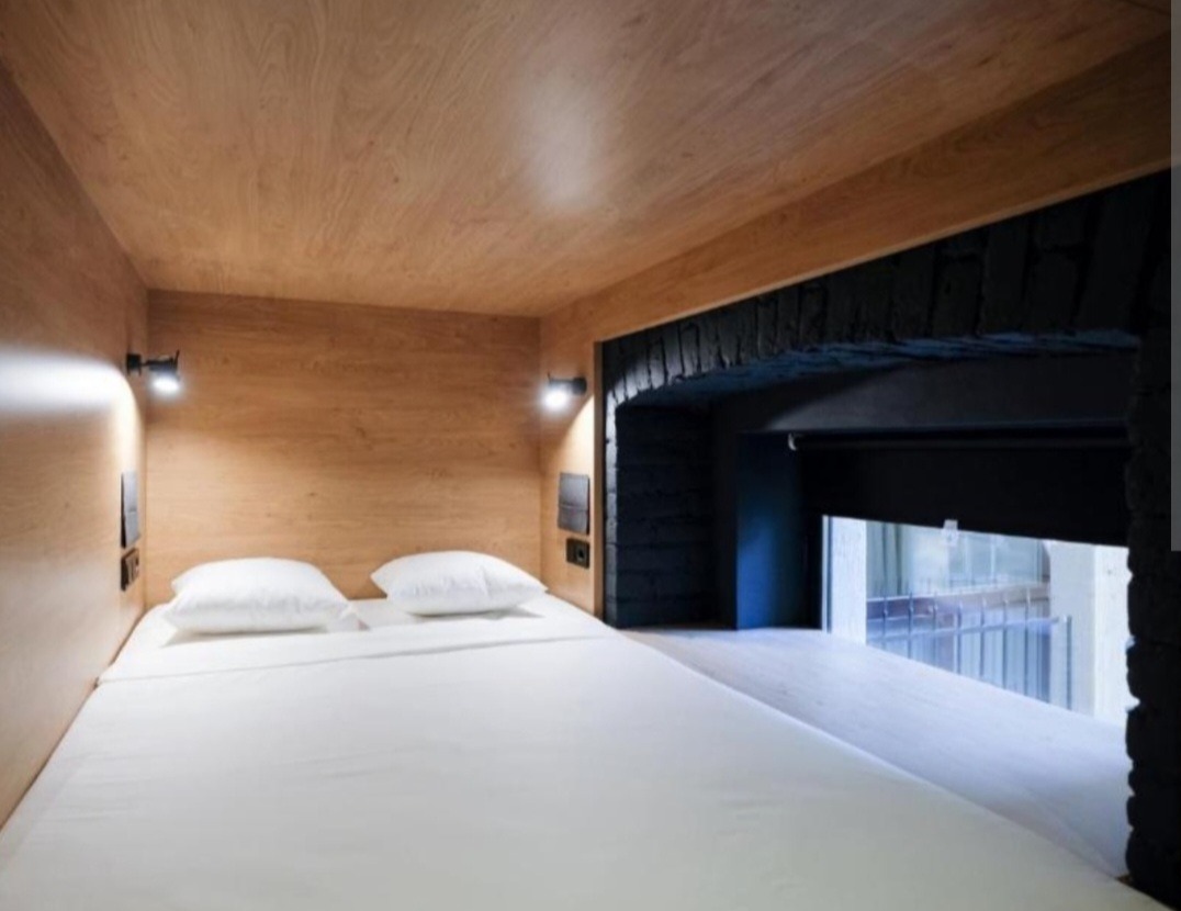 Minimalistyczna sypialnia w kapsule z drewnianym wykończeniem, białymi poduszkami i oknem z widokiem na miasto.