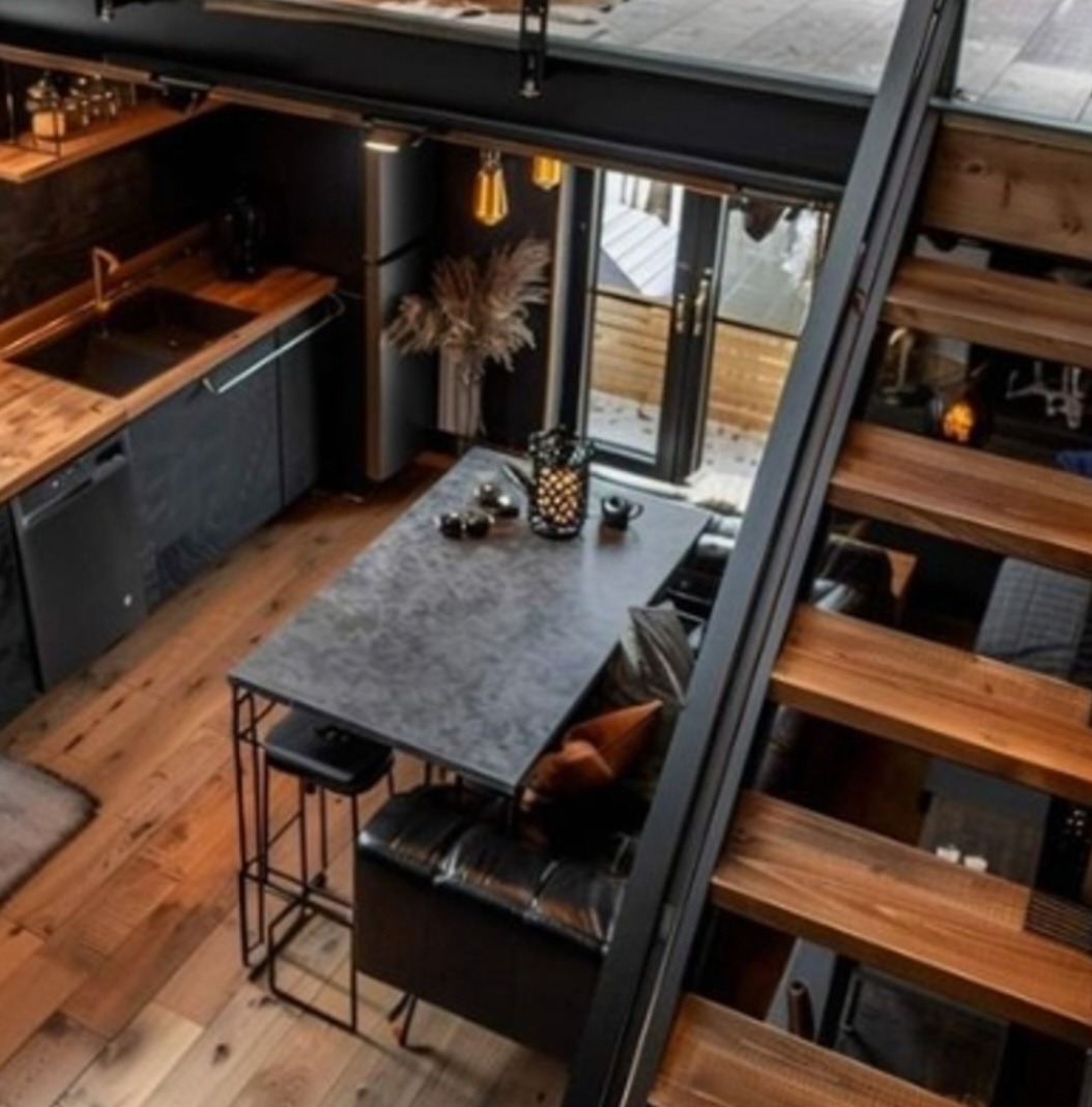 Loftowe wnętrze z drewnianą podłogą, czarną zabudową kuchenną i schodami z drewnianymi stopniami, widok z góry na stół z krzesłem i kanapą.