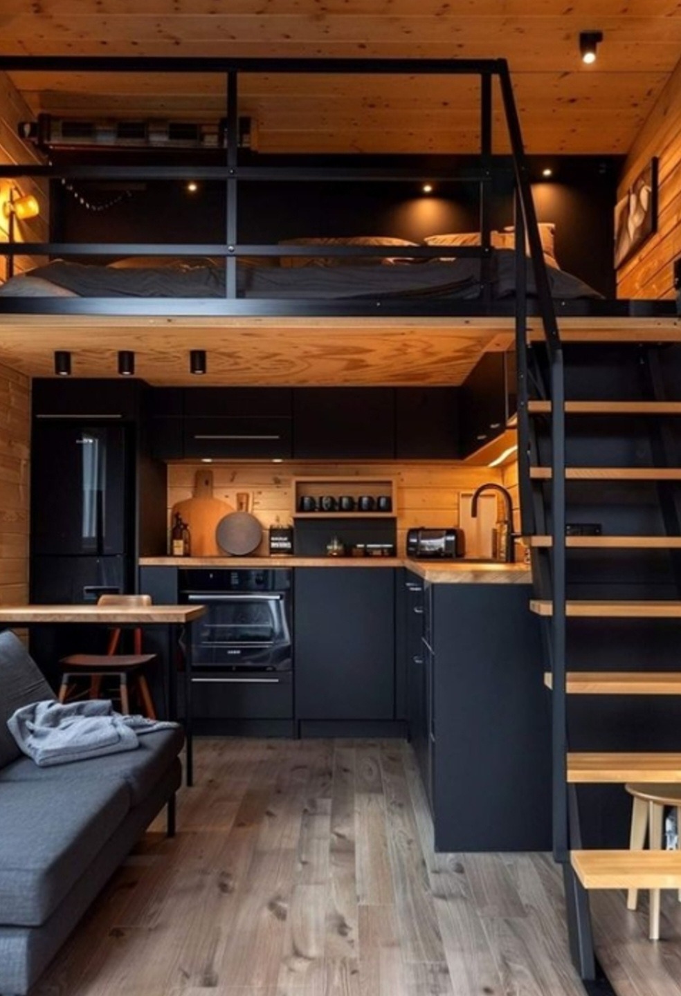 Małe, ale funkcjonalne wnętrze z antresolą sypialnianą, czarną zabudową kuchenną na tle drewnianej ściany, drewnianymi schodami i szarą sofą, oświetlone punktowymi lampami.