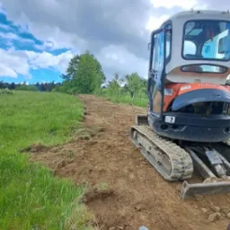 Mini koparka Kubota na terenie zielonym, przygotowująca grunt pod ścieżkę, z widocznymi śladami pracy i kubkiem na ziemi.