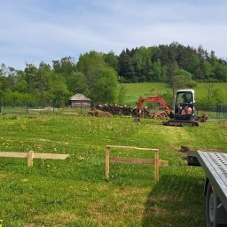 Koparka Arka - Mini koparka w trakcie pracy na zielonej działce, widoczne drewniane szalunki, ułożone drewno, w tle dom i las.