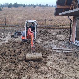 Koparka Arka - Mini koparka w akcji przy wykopie obok budynku w stanie surowym otwartym. Widoczne rury drenażowe i hałda ziemi. Praca w toku, pochmurny dzień.