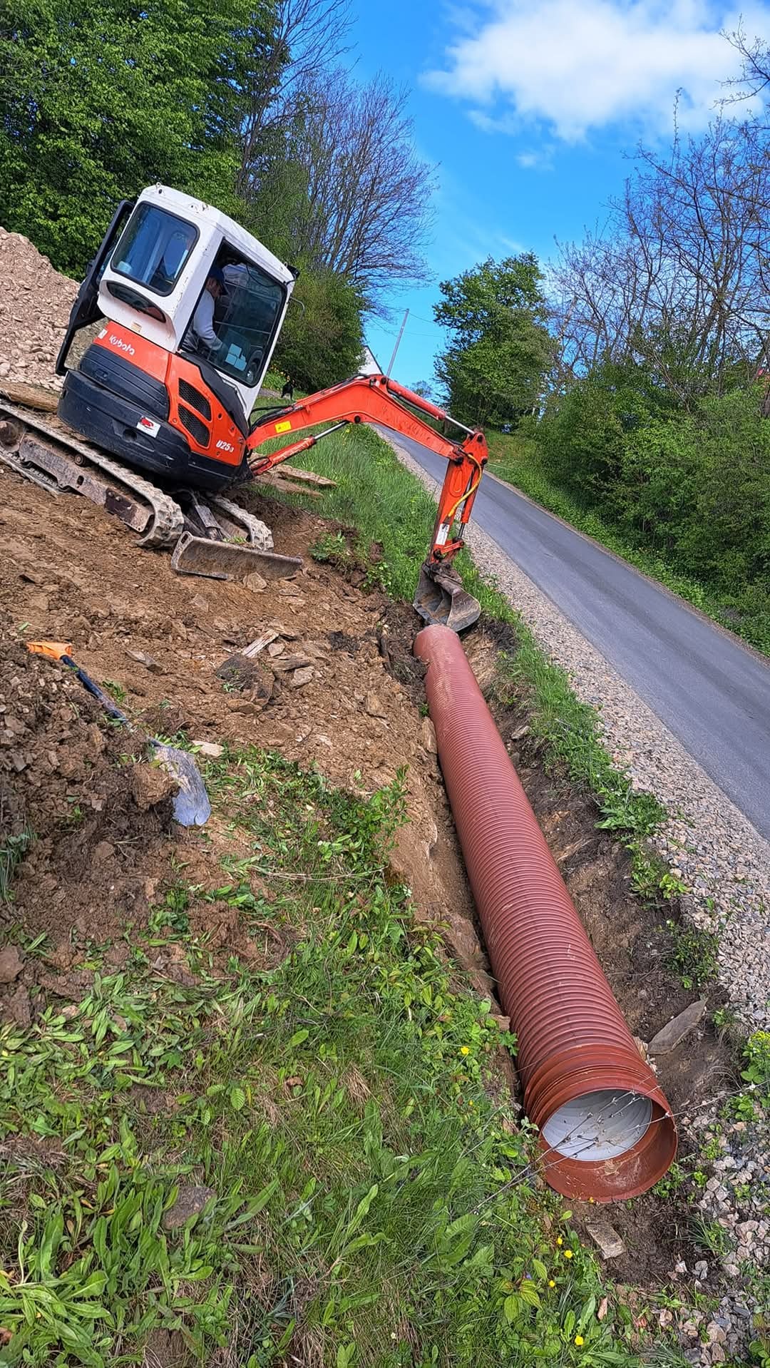 Pomarańczowa rura drenażowa w wykopie obok drogi, z koparką Kubota w tle, widok z góry na zbocze z trawą i ziemią.