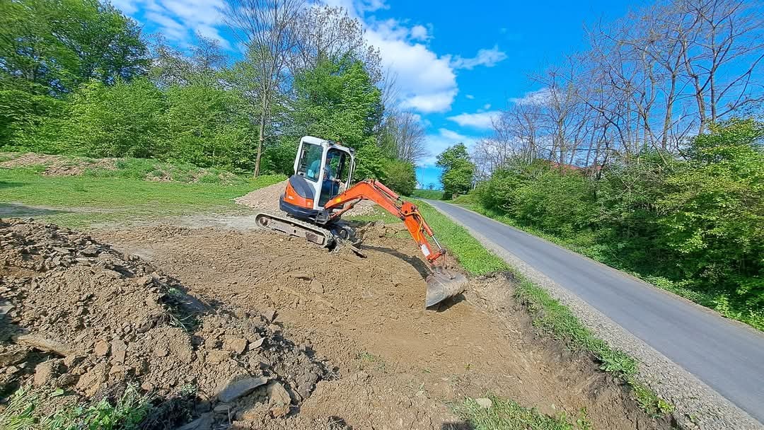Pomarańczowo-biała mini koparka wykonuje wykop na zboczu, obok asfaltowej drogi, w tle zielone drzewa i błękitne niebo z chmurami.