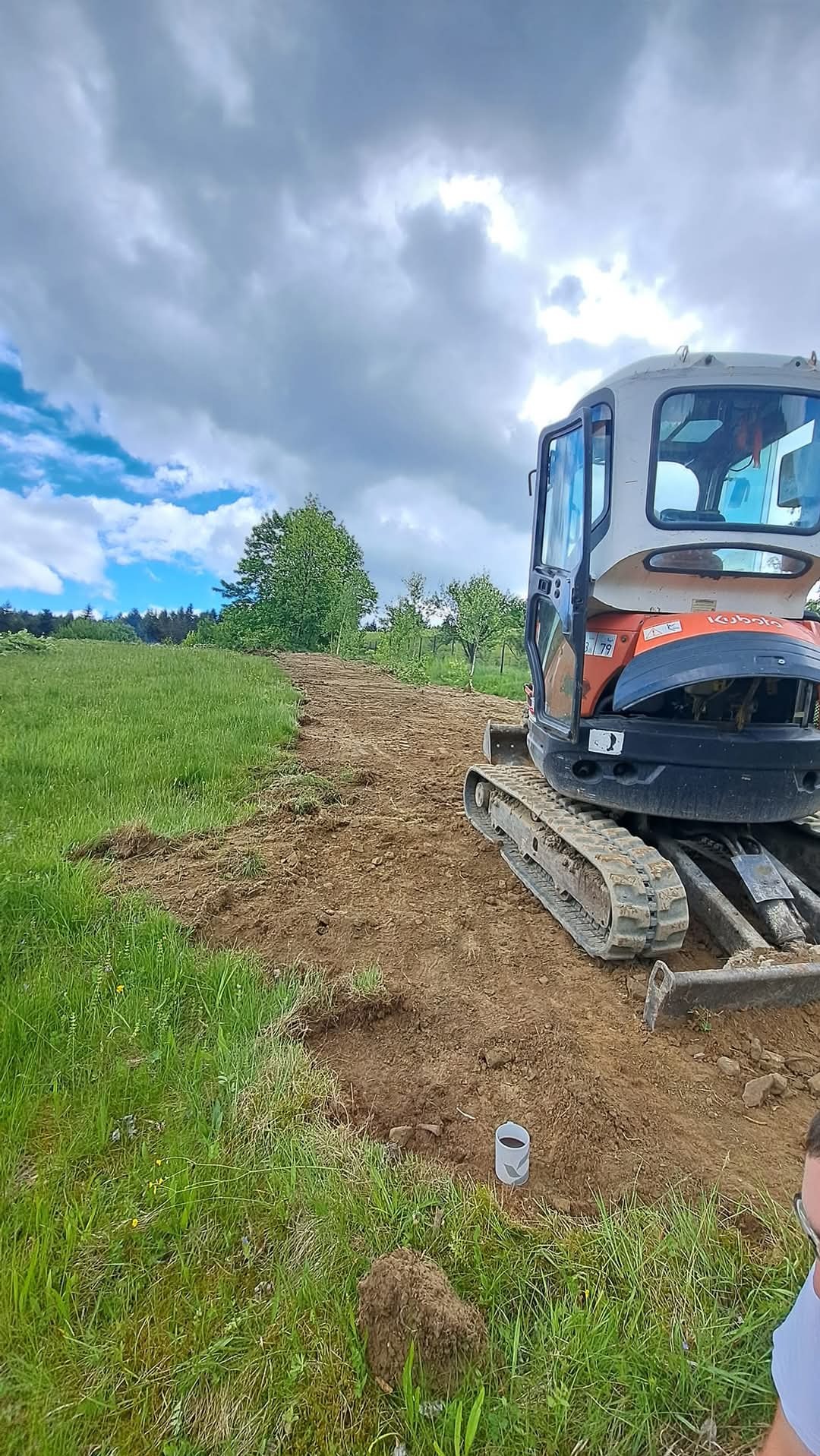 Mini koparka Kubota na terenie zielonym, przygotowująca grunt pod ścieżkę, z widocznymi śladami pracy i kubkiem na ziemi.