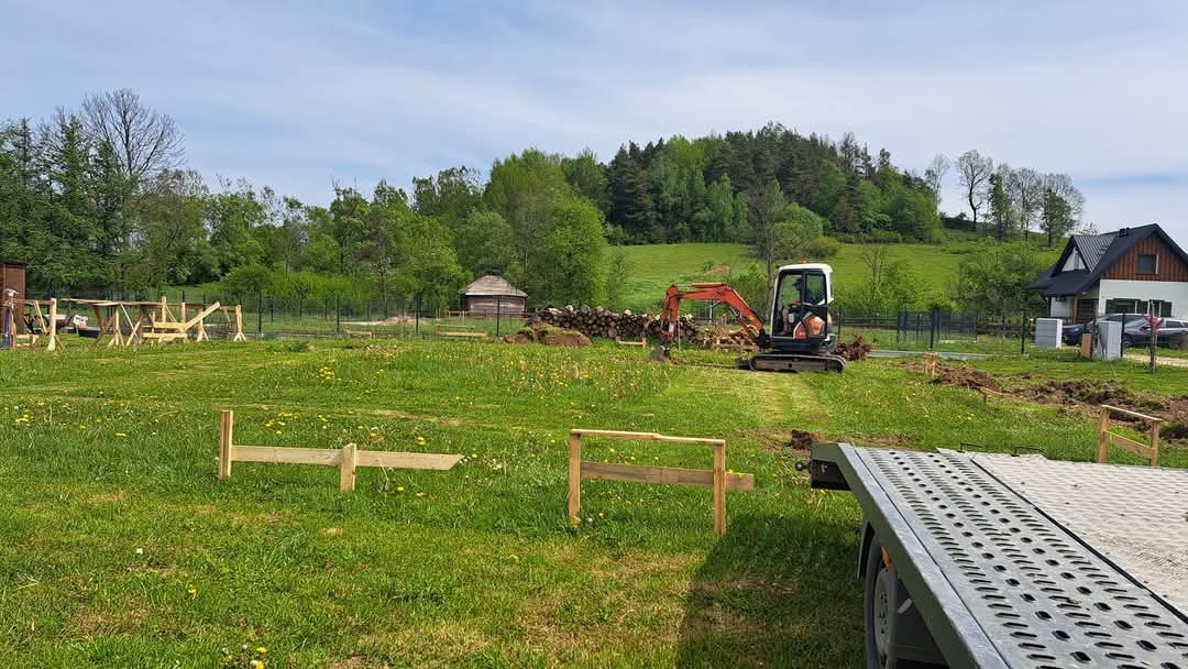 Mini koparka w trakcie pracy na zielonej działce, widoczne drewniane szalunki, ułożone drewno, w tle dom i las.
