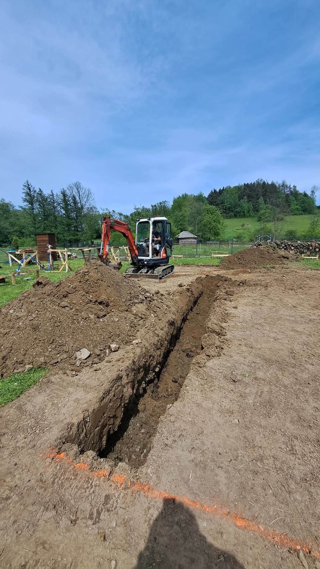 Mini koparka przy wykopie pod przyszły fundament, pomarańczowe oznaczenia wyznaczają linię, w tle widać drewniane konstrukcje i zalesione wzgórze.