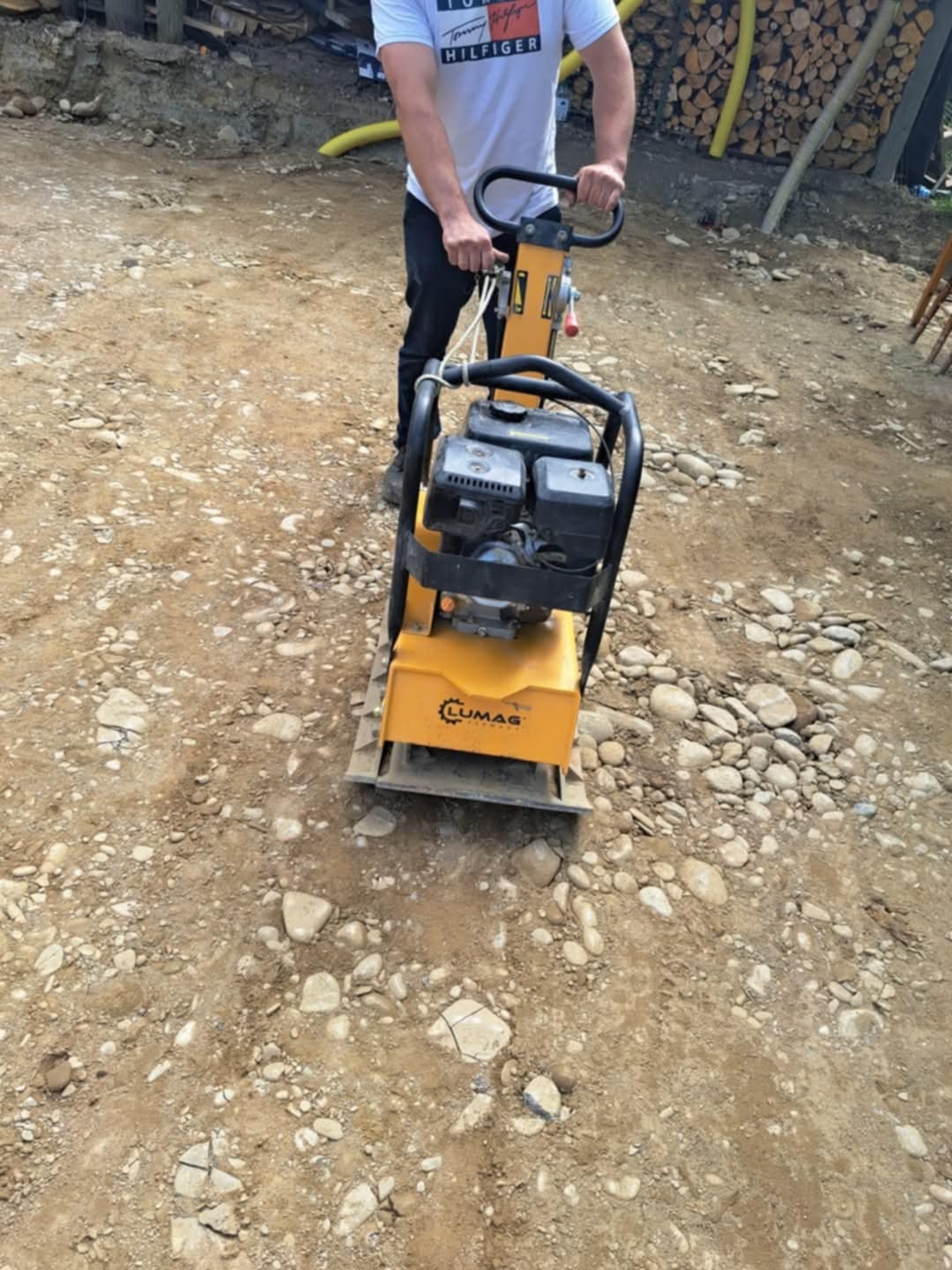 Pracownik ubija ziemię zagęszczarką LUMAG na terenie budowy. W tle widoczne drewno opałowe i żółty wąż. Teren przygotowywany pod dalsze prace.