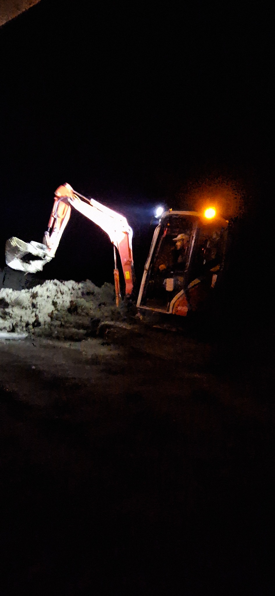 Oświetlony pomarańczowym światłem reflektorów, pracujący w nocy na placu budowy, mały, czerwony traktor z łyżką nabiera ziemię. Widoczny operator w kabinie.