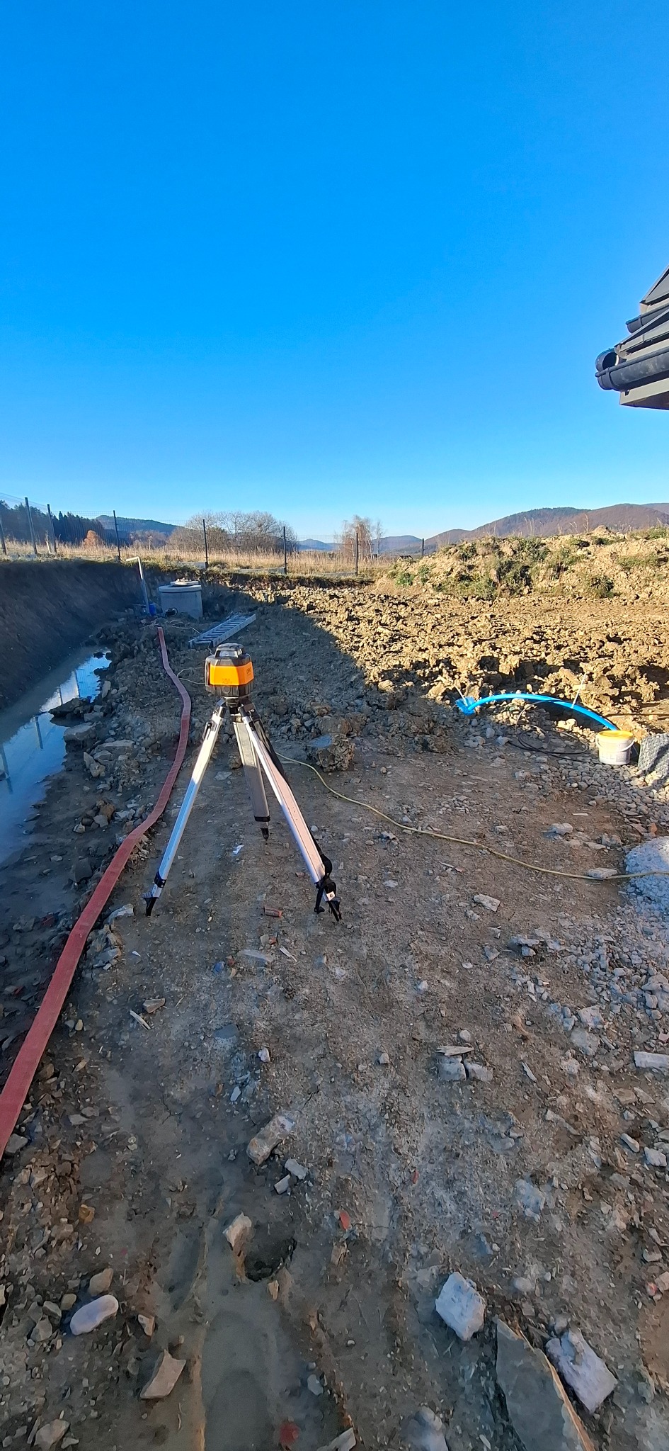 Prace ziemne w terenie: wykop z wodą, rury, poziomica laserowa na statywie. W tle widoczne wzgórza i błękitne niebo. Prace przygotowawcze do budowy.