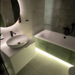 Nowoczesna łazienka z okrągłym lustrem podświetlanym LED, owalną umywalką nablatową i podświetlaną wanną, wykończona płytkami w odcieniach szarości i zieleni.