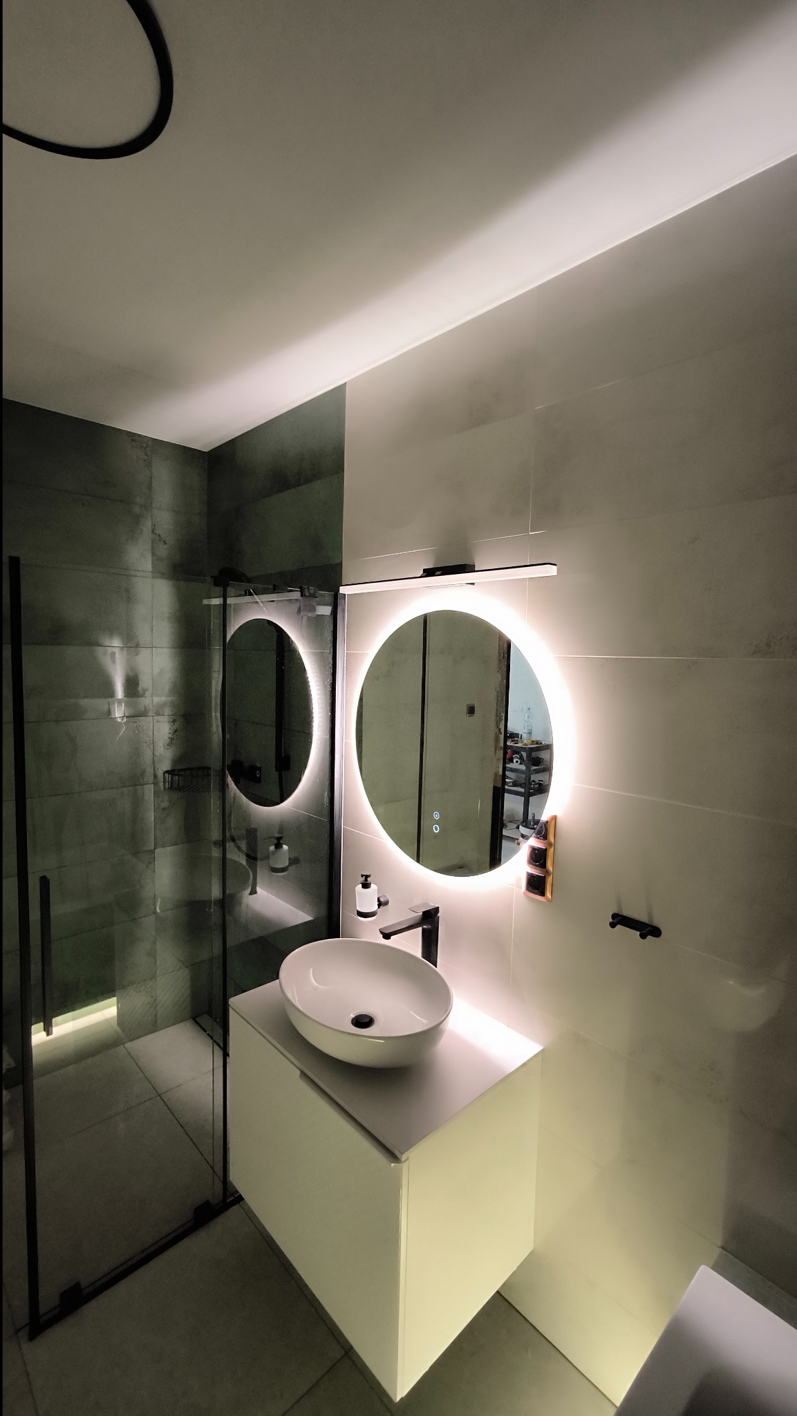 Nowoczesna łazienka z kabiną prysznicową typu walk-in, okrągłym lustrem z podświetleniem LED, białą umywalką nablatową i czarną armaturą.