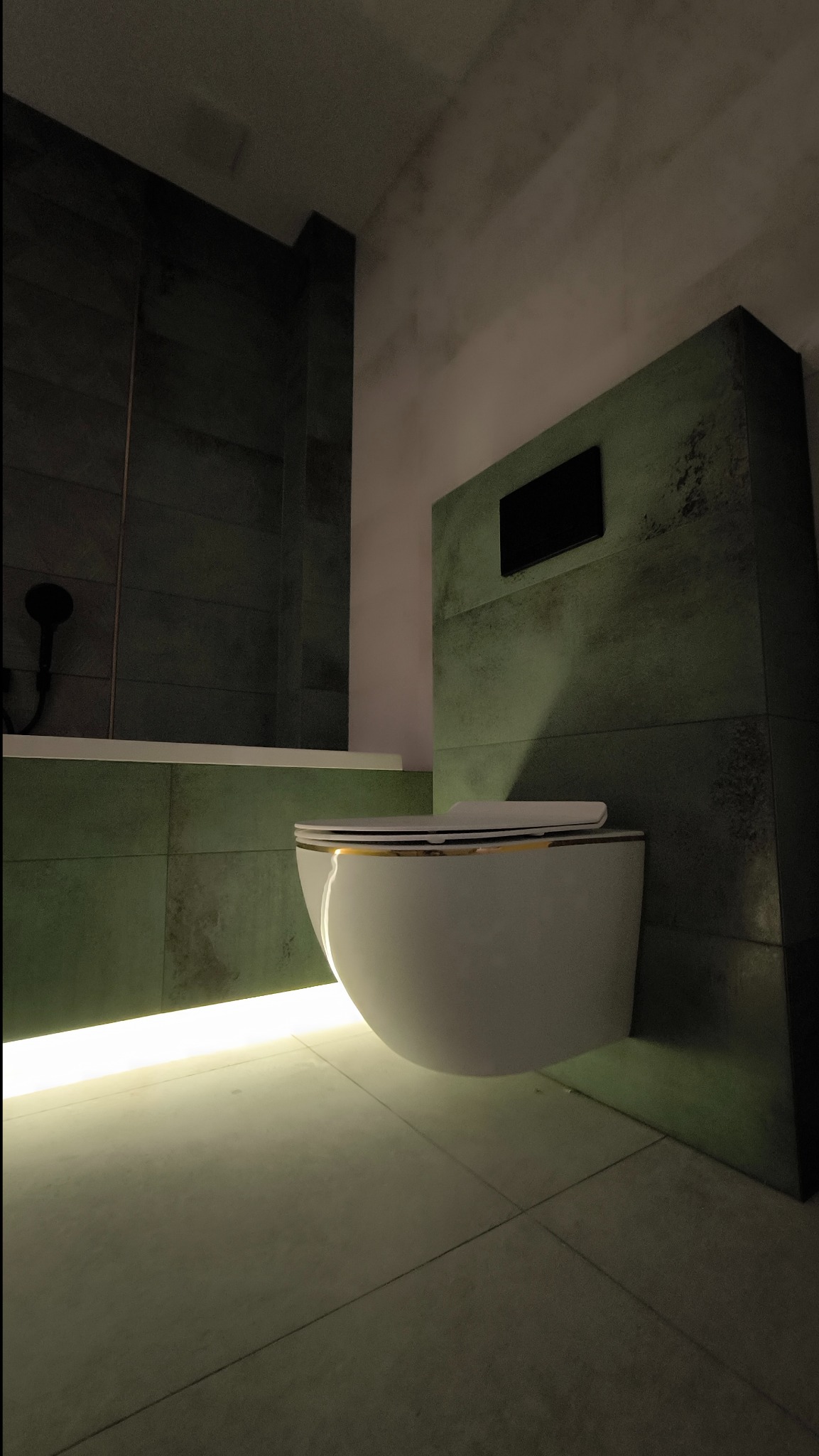 Nowoczesna toaleta wisząca z podświetleniem LED u dołu, zamontowana na ścianie wyłożonej płytkami w odcieniach zieleni, z widoczną wanną w tle.