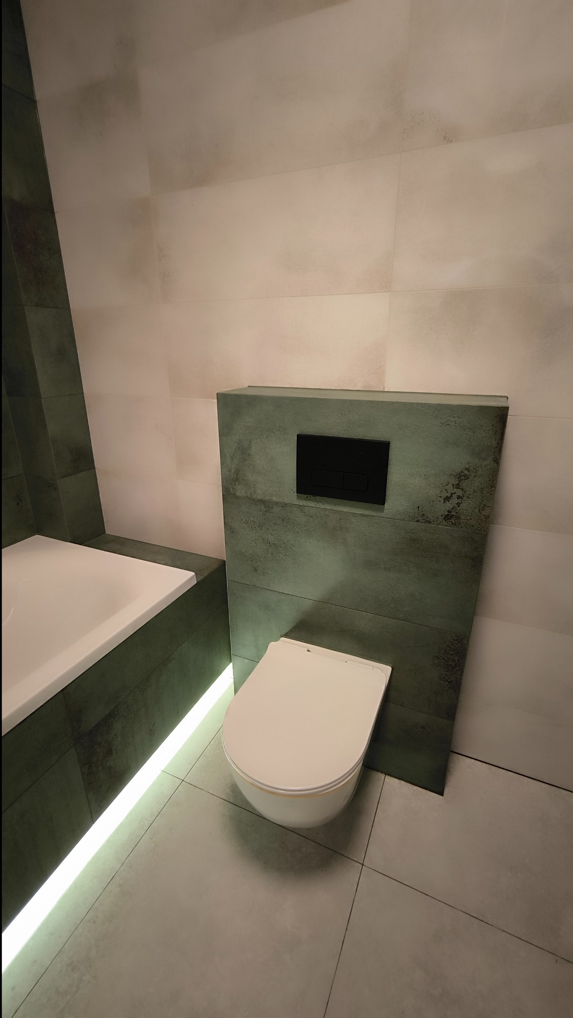 Nowoczesna toaleta podwieszana z czarnym panelem sterującym na tle płytek imitujących beton, podświetlana listwa LED pod wanną, szare płytki podłogowe.