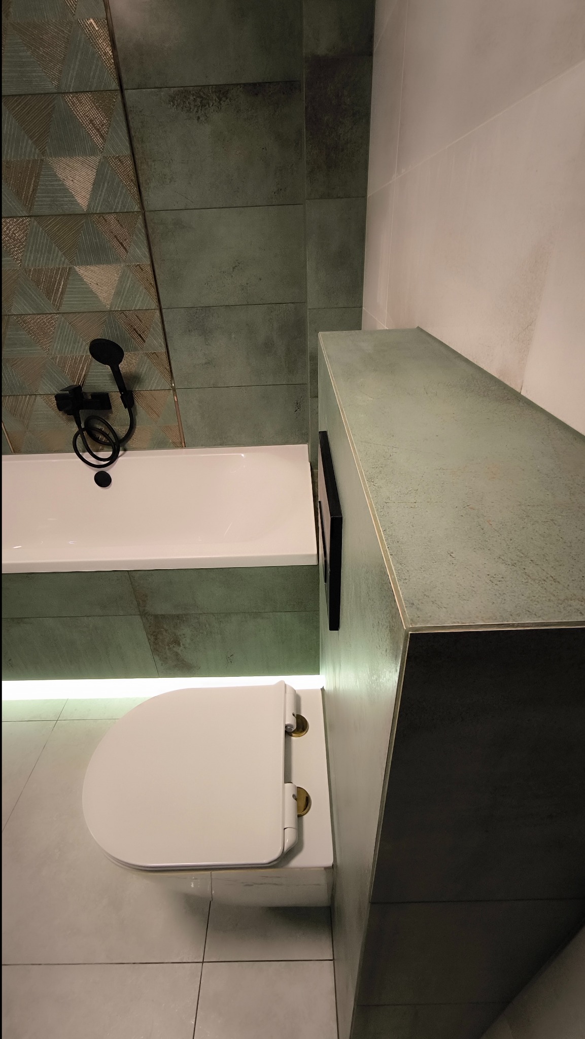 Nowoczesna łazienka z zielonymi płytkami o zróżnicowanej fakturze, białą wanną z czarną armaturą, podświetleniem LED i podwieszaną toaletą z deską wolnoopadającą, widok z góry.