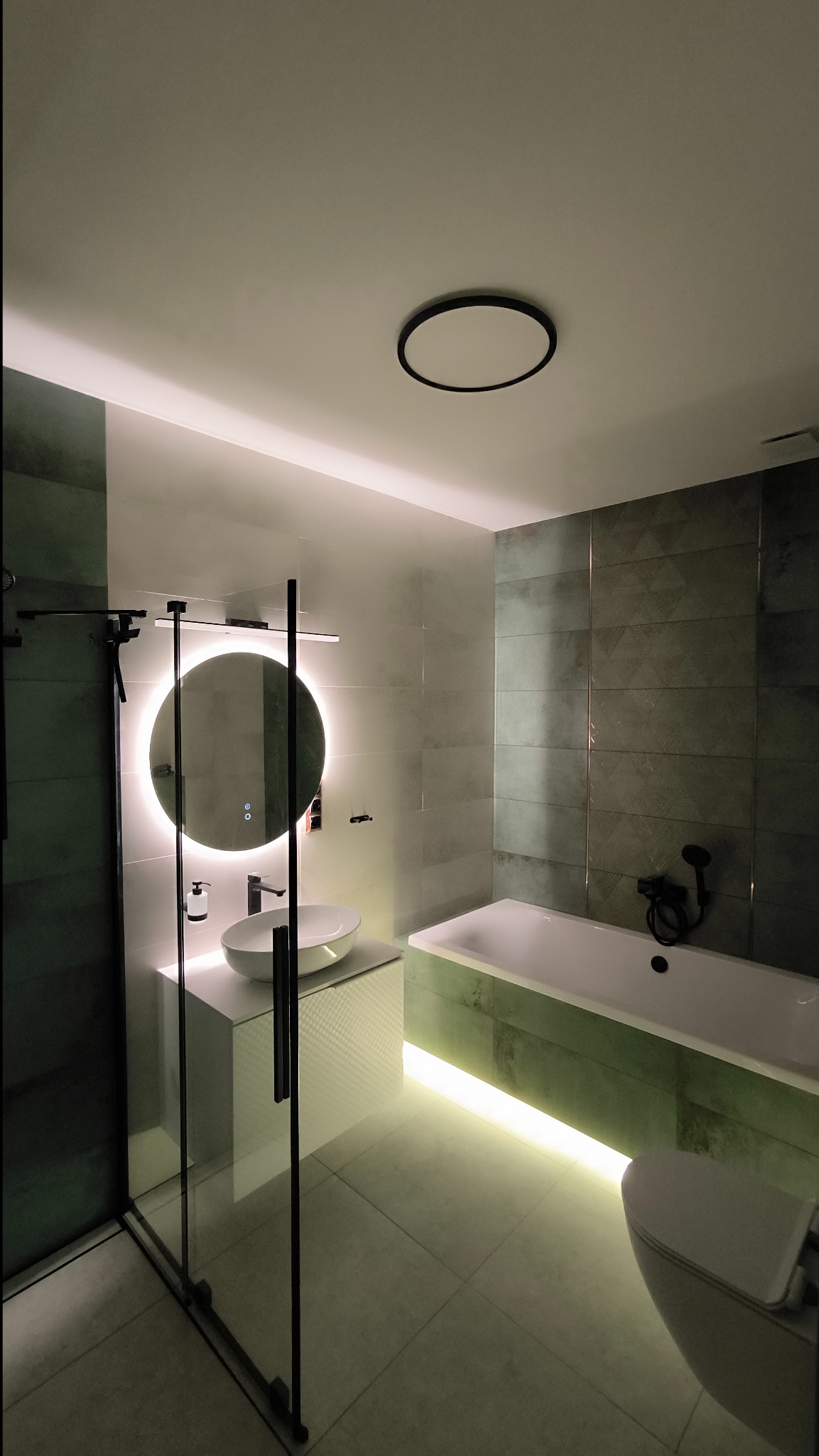 Nowoczesna łazienka z podświetlaną wanną, okrągłym lustrem z podświetleniem LED, kabiną prysznicową ze szklanymi drzwiami z czarnymi ramami i umywalką nablatową.