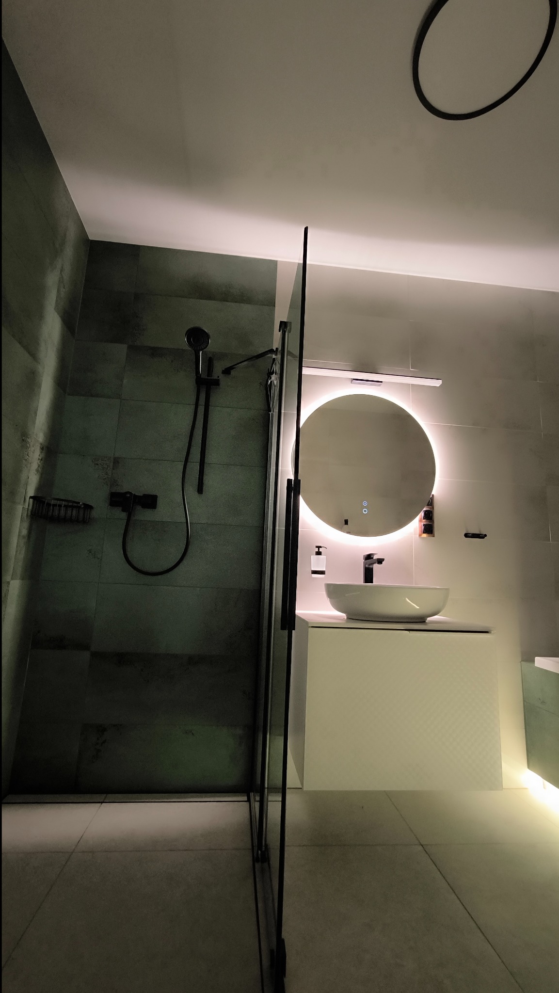 Nowoczesna łazienka z kabiną prysznicową typu walk-in, czarną armaturą i okrągłym lustrem z podświetleniem LED, widok z dołu.