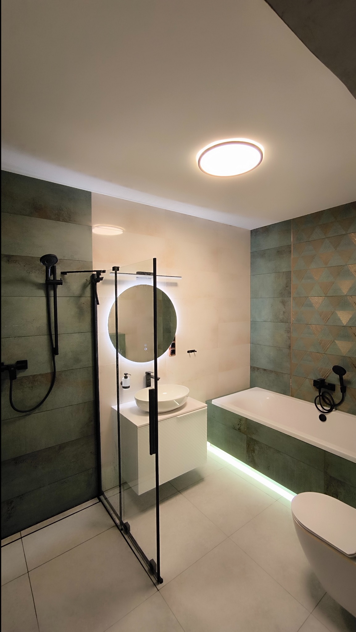 Nowoczesna łazienka z zielonymi płytkami o geometrycznym wzorze, kabiną prysznicową ze szkła hartowanego z czarnymi okuciami, okrągłym lustrem z podświetleniem LED, białą umywalką nablatową...