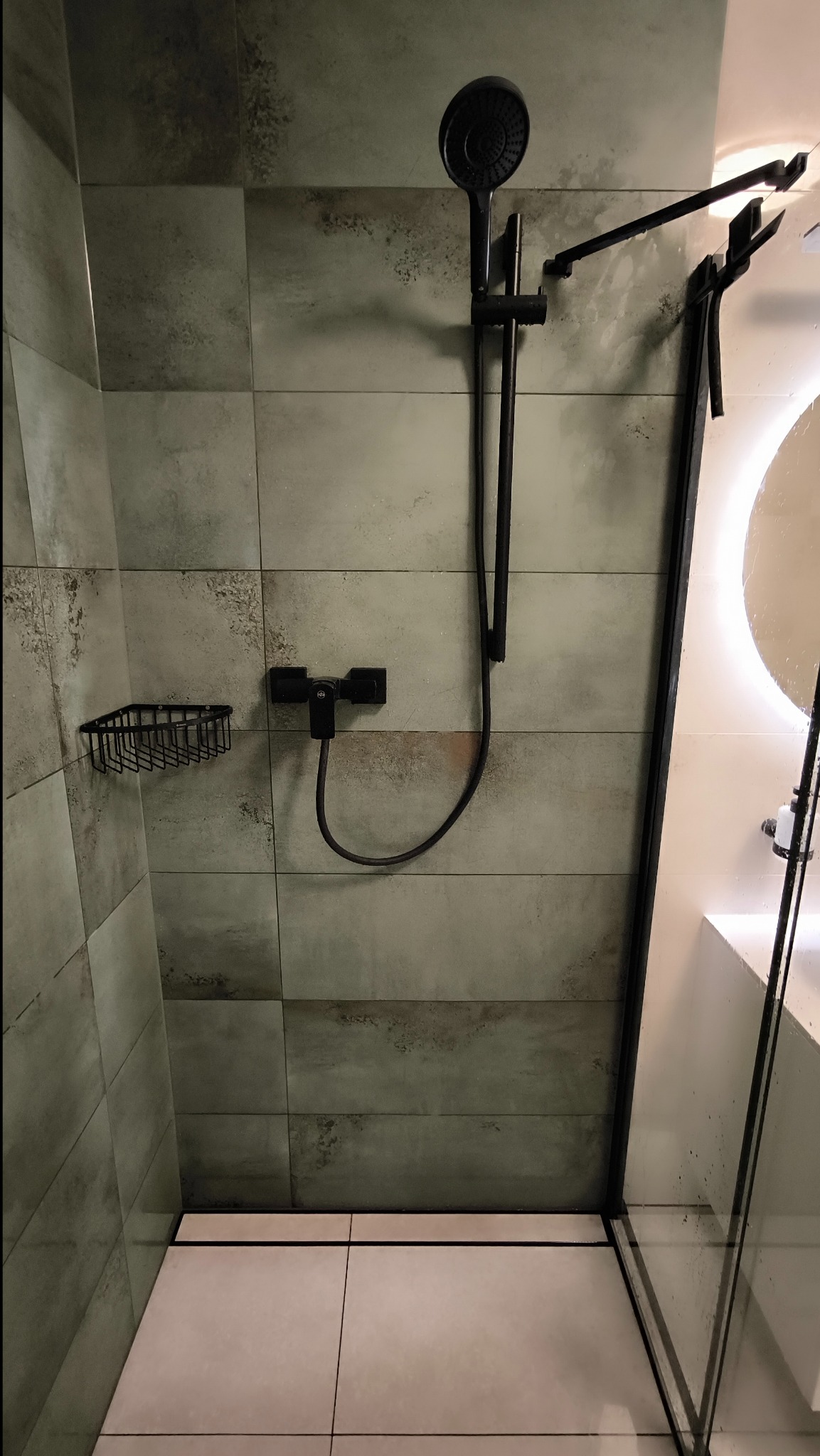 Nowoczesna kabina prysznicowa z czarną armaturą, płytki ścienne w odcieniach szarości i zieleni z delikatną teksturą, biała podłoga z odpływem liniowym, czarny koszyk na kosmetyki.