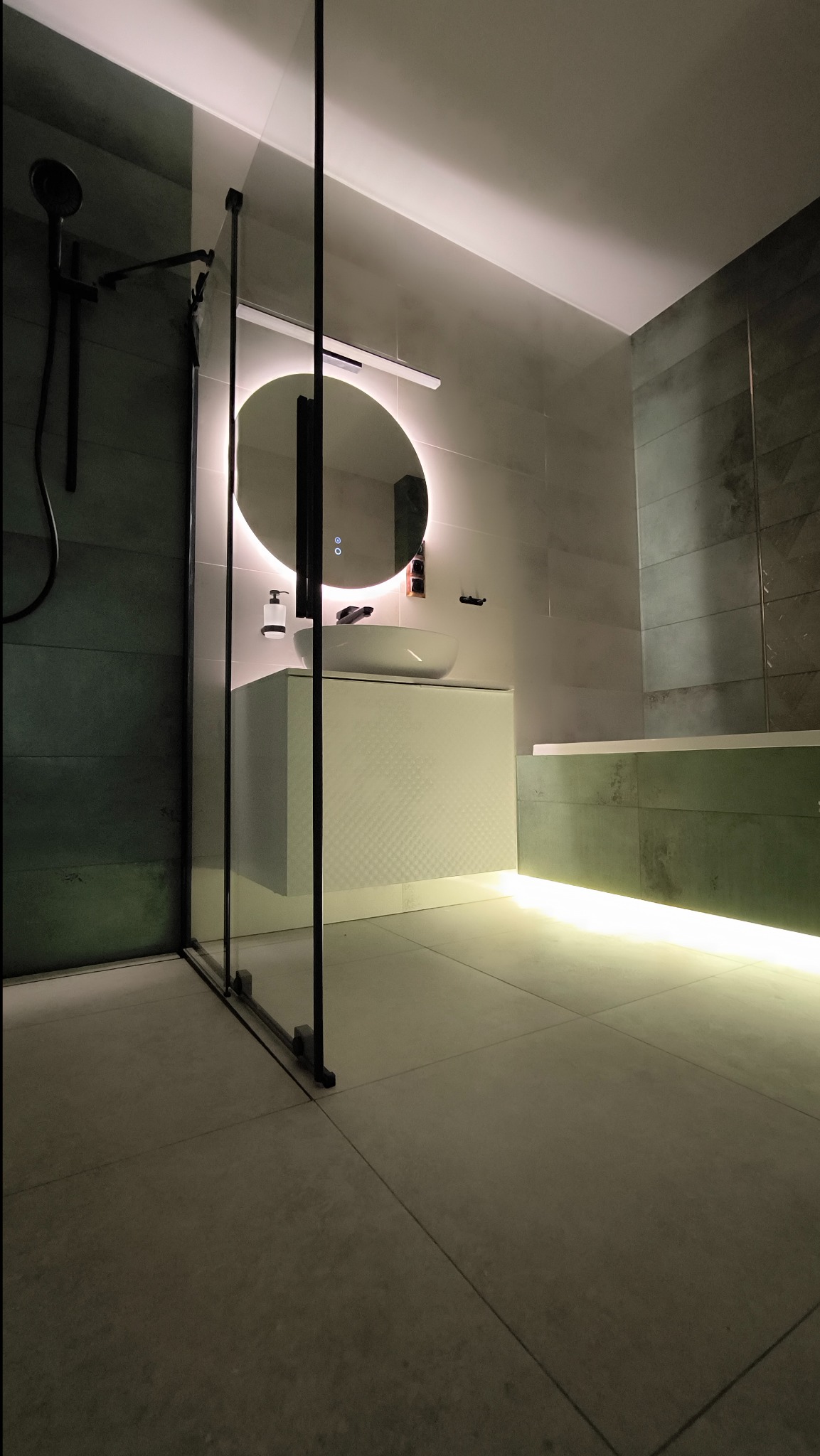Nowoczesna łazienka z podświetlanym lustrem, szklaną kabiną prysznicową i podświetloną wanną, utrzymana w odcieniach szarości i zieleni.