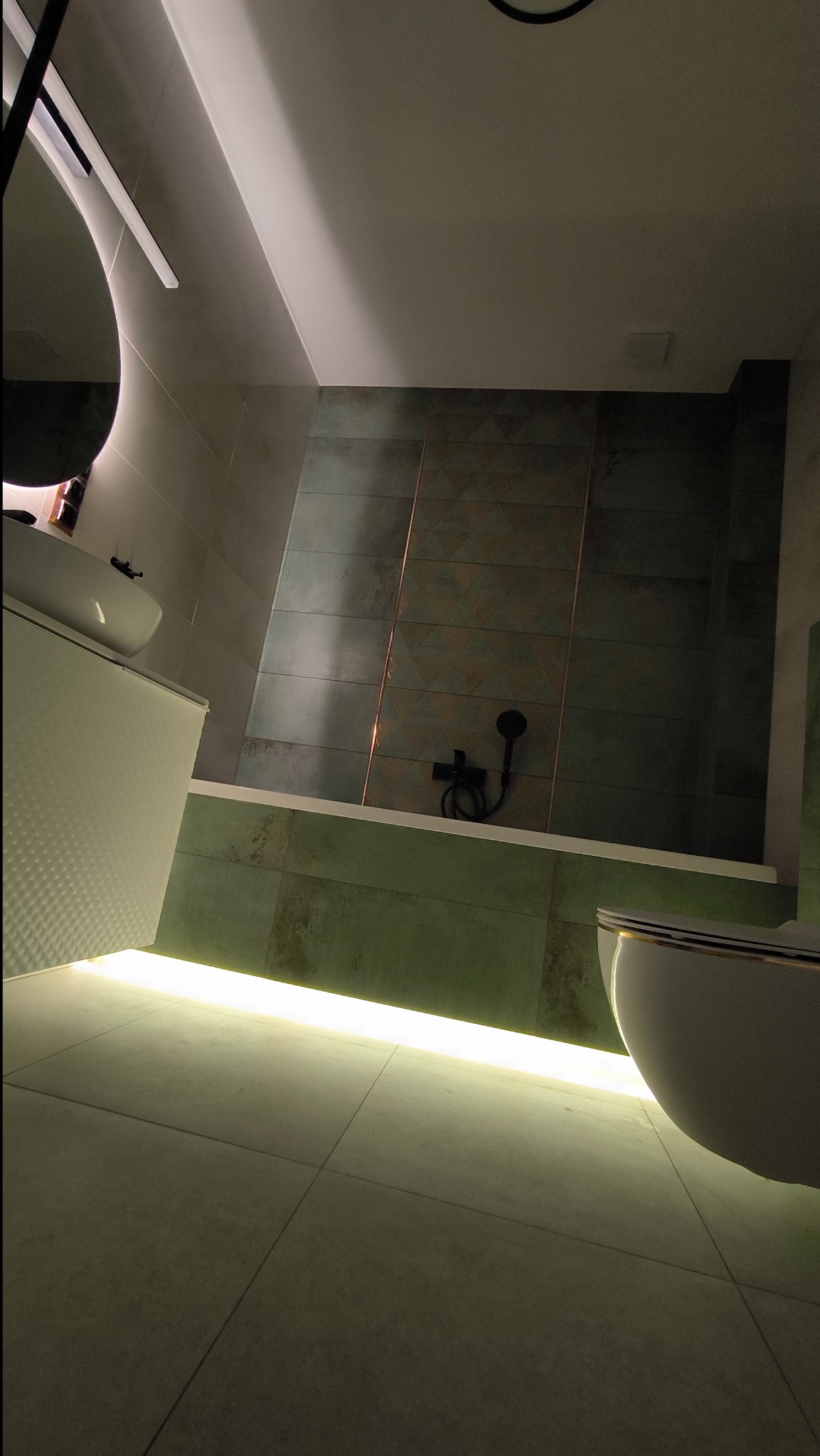 Nowoczesna łazienka z podświetloną wanną, zielonymi płytkami z geometrycznym wzorem, czarną armaturą i okrągłym lustrem z podświetleniem LED.