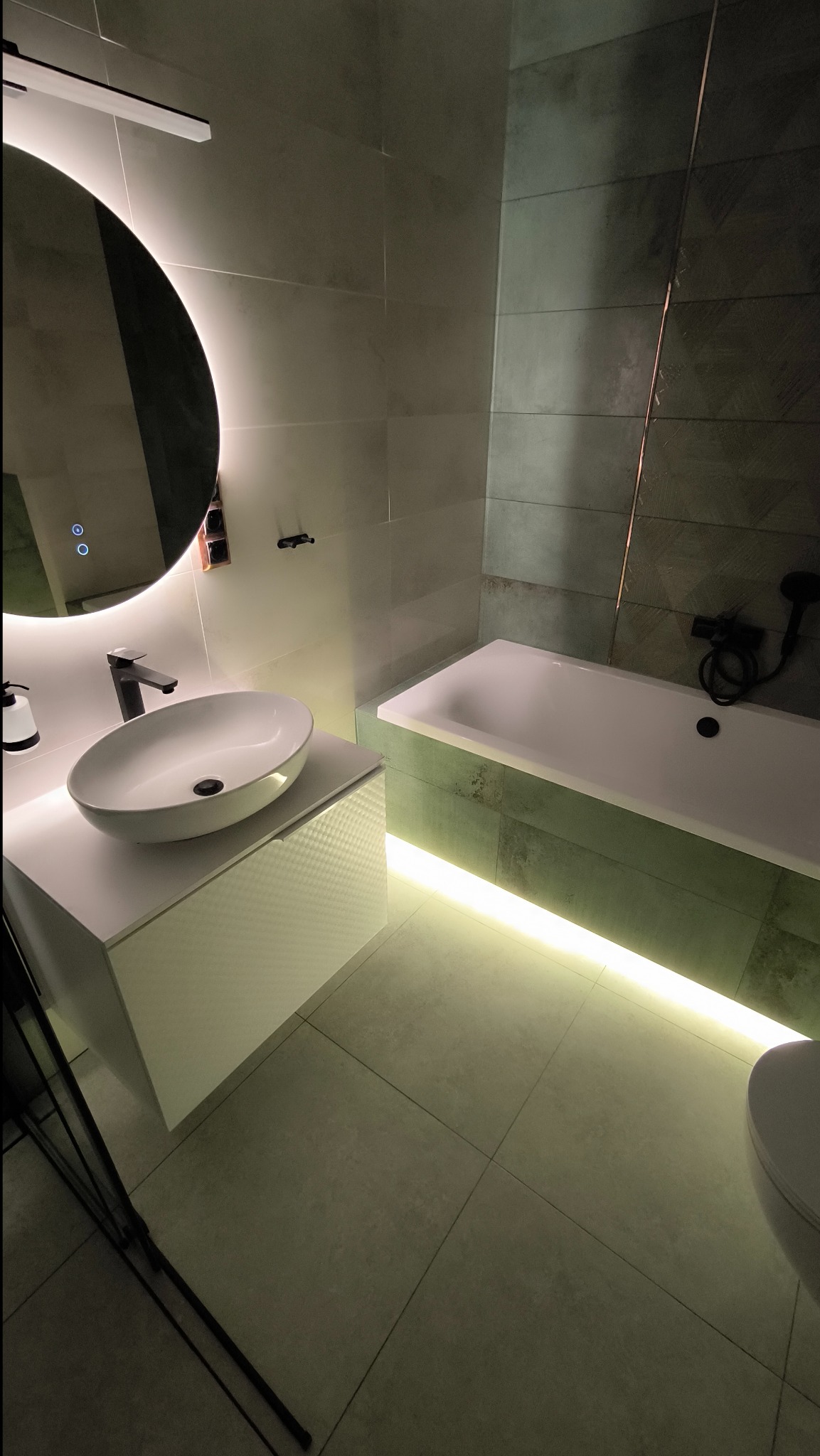 Nowoczesna łazienka z okrągłym lustrem podświetlanym LED, owalną umywalką nablatową i podświetlaną wanną, wykończona płytkami w odcieniach szarości i zieleni.
