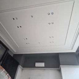 KOLIN BUD - Sufit z dekoracyjną sztukaterią w ramce i punktowym oświetleniem, ściany częściowo pomalowane na szaro z widocznym klimatyzatorem, dolna część ścian przygotowana do malowania.