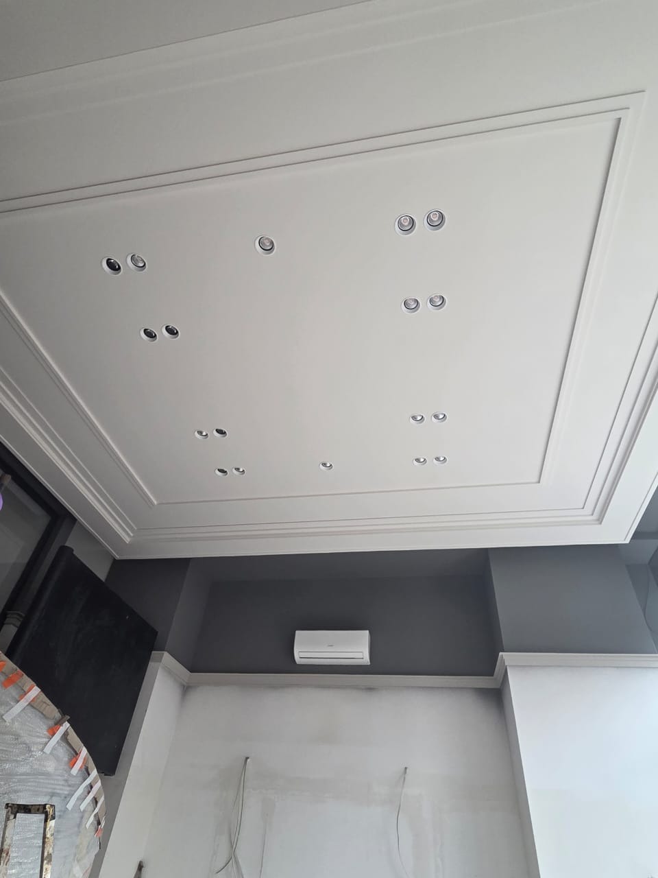 Sufit z dekoracyjną sztukaterią w ramce i punktowym oświetleniem, ściany częściowo pomalowane na szaro z widocznym klimatyzatorem, dolna część ścian przygotowana do malowania.