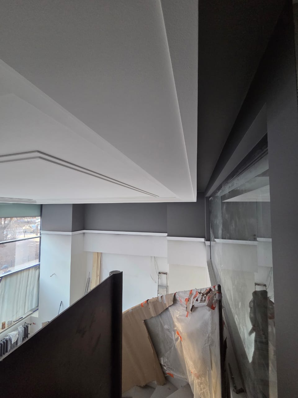 Wnętrze biura w trakcie odświeżania: białe ściany częściowo pomalowane na szaro, widoczne zabezpieczenia i drabina, sufit z ozdobnymi listwami.