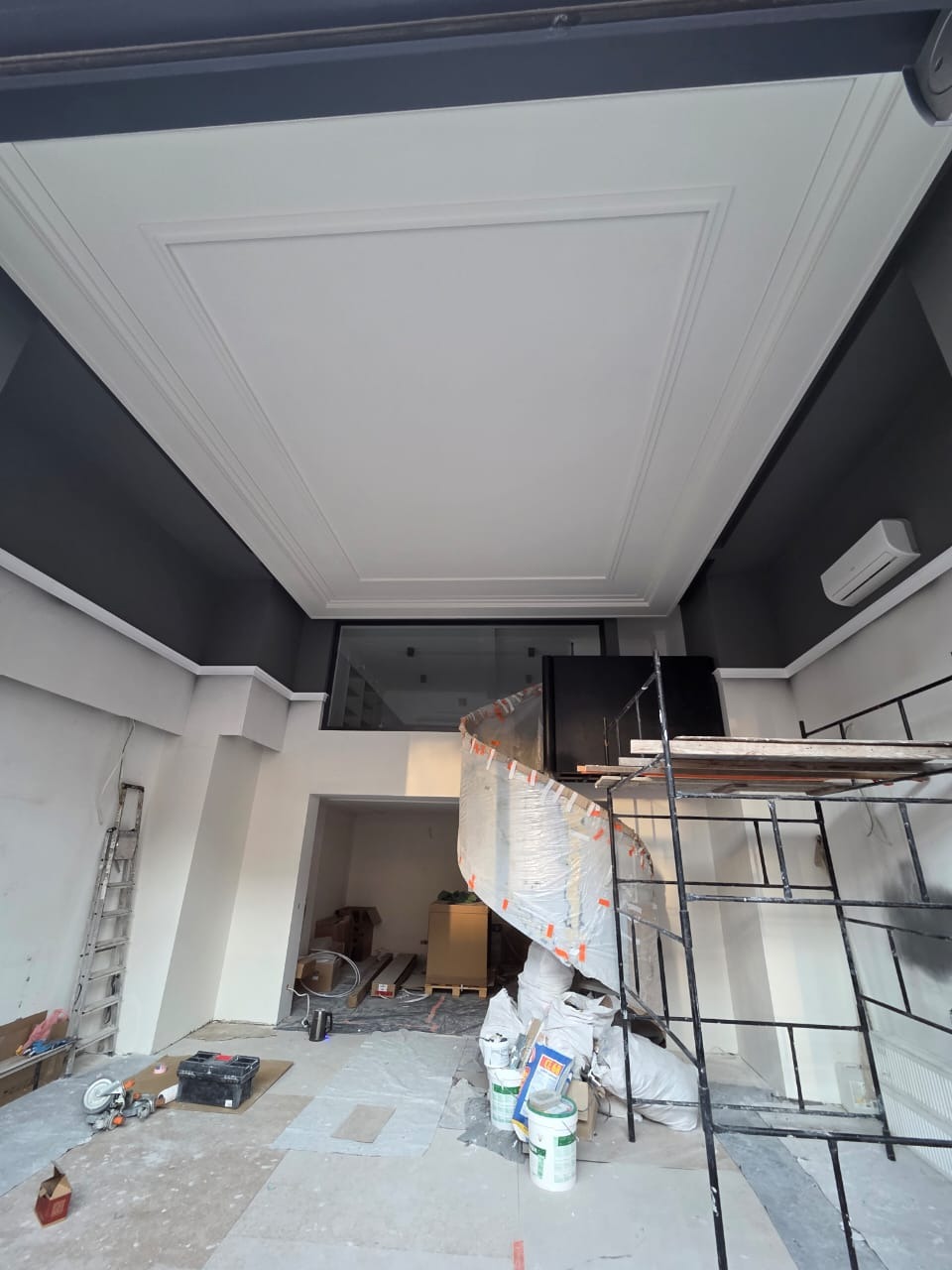 Biały, kasetonowy sufit z dekoracyjnymi listwami w remontowanym pomieszczeniu biurowym, widoczne rusztowanie, drabina i narzędzia malarskie.