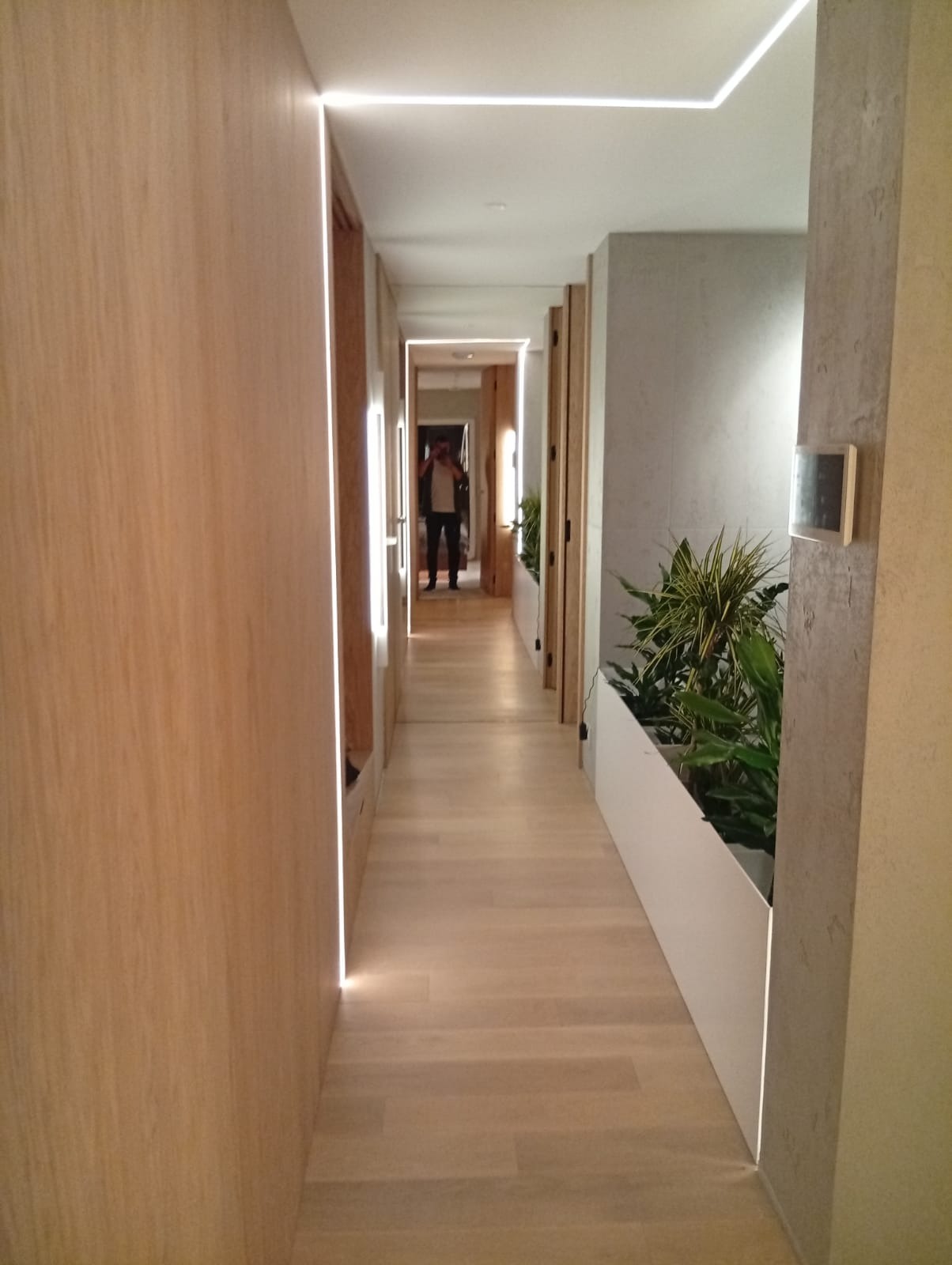 Długi korytarz z drewnianą podłogą i oświetleniem LED wzdłuż ścian, zakończony lustrem, w którym odbija się osoba.