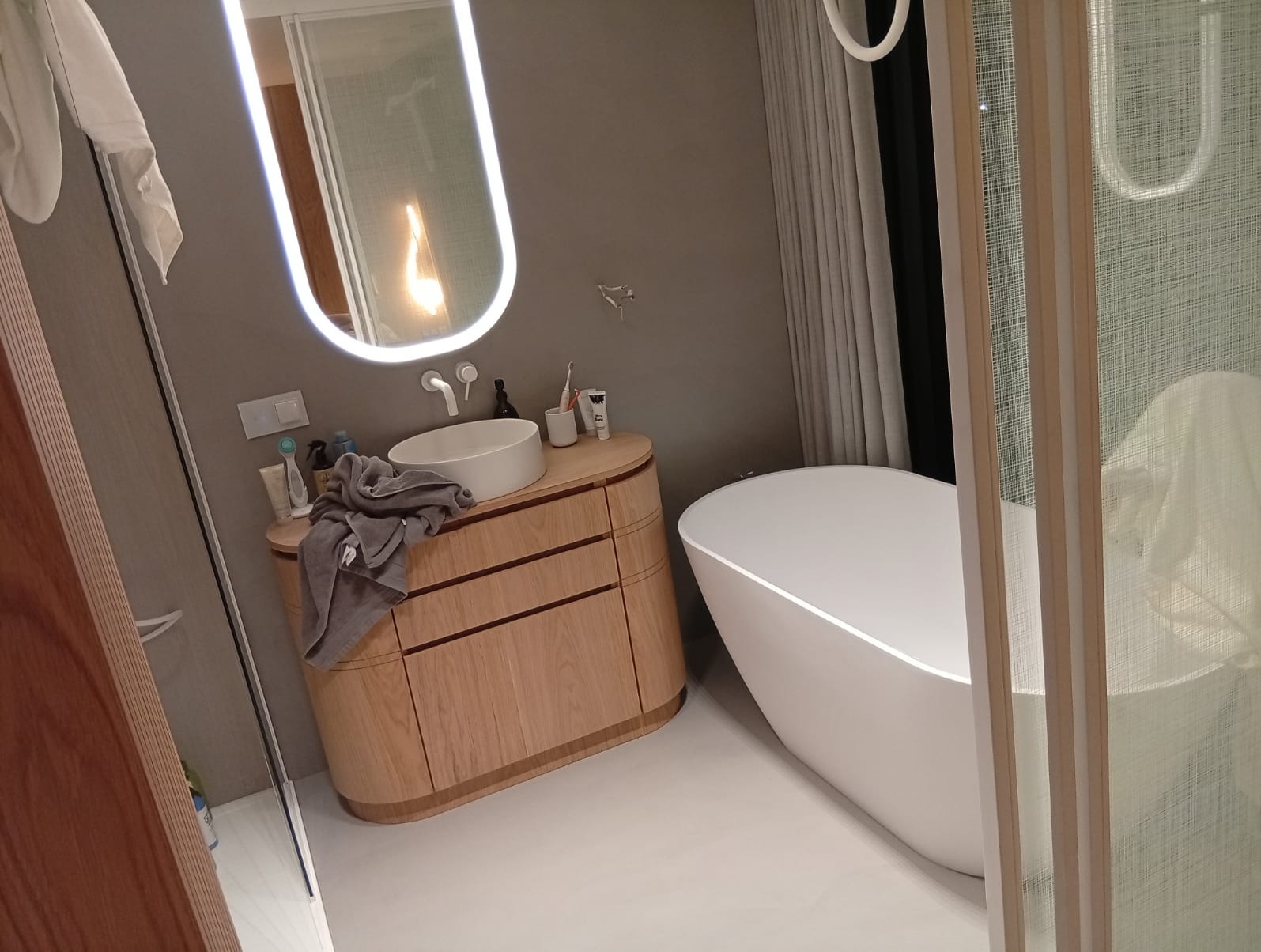 Nowoczesna łazienka z wolnostojącą wanną, umywalką nablatową na drewnianej szafce z szufladami i podświetlanym lustrem owalnym, szary ręcznik leży na blacie.