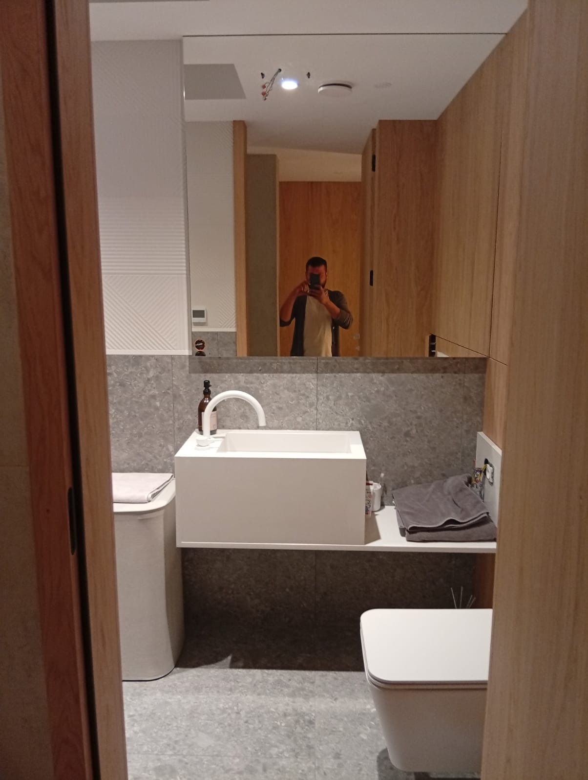 Nowoczesna łazienka z umywalką nablatową, szarą płytką i drewnianymi panelami, widoczne odbicie fotografa w lustrze.