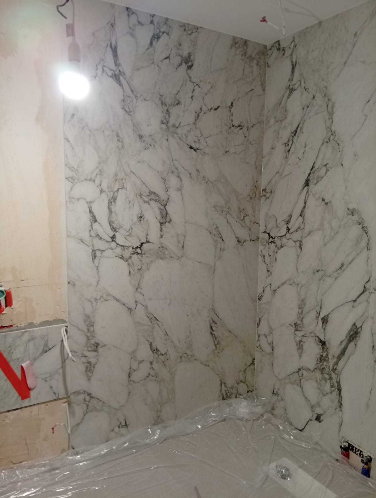 Ściany łazienki wyłożone białymi płytami imitującymi marmur, zabezpieczone folią podczas prac wykończeniowych, widoczna surowa ściana z instalacjami.