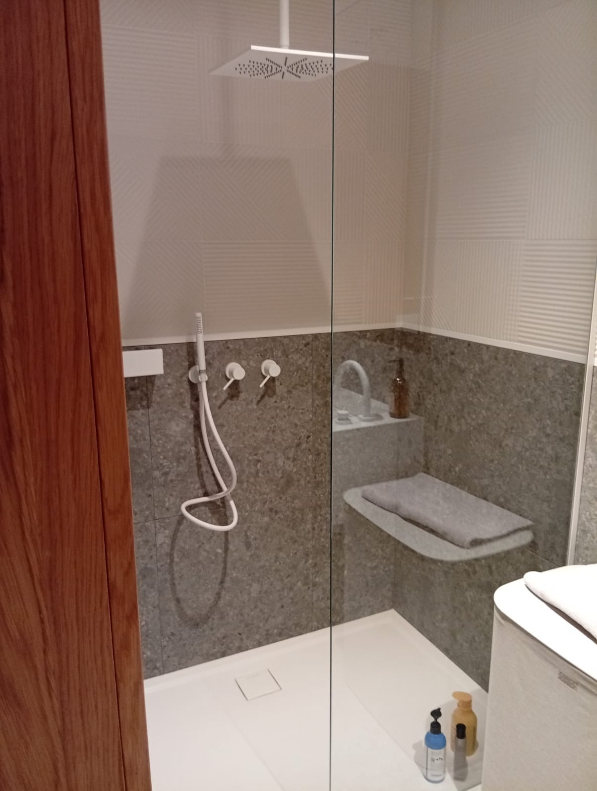 Nowoczesna kabina prysznicowa z deszczownicą, szklaną ścianą, szarą mozaiką na ścianach, białą półką z ręcznikiem i minimalistyczną armaturą.